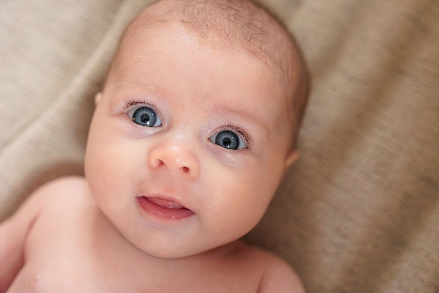 Smiling baby with bright eyes lying on soft beige fabric showing a happy expression.