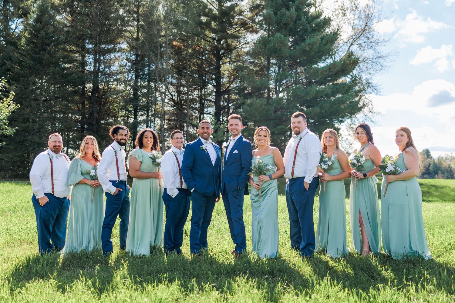 Large wedding party in mint green dresses and navy blue attire poses together on grassy field.