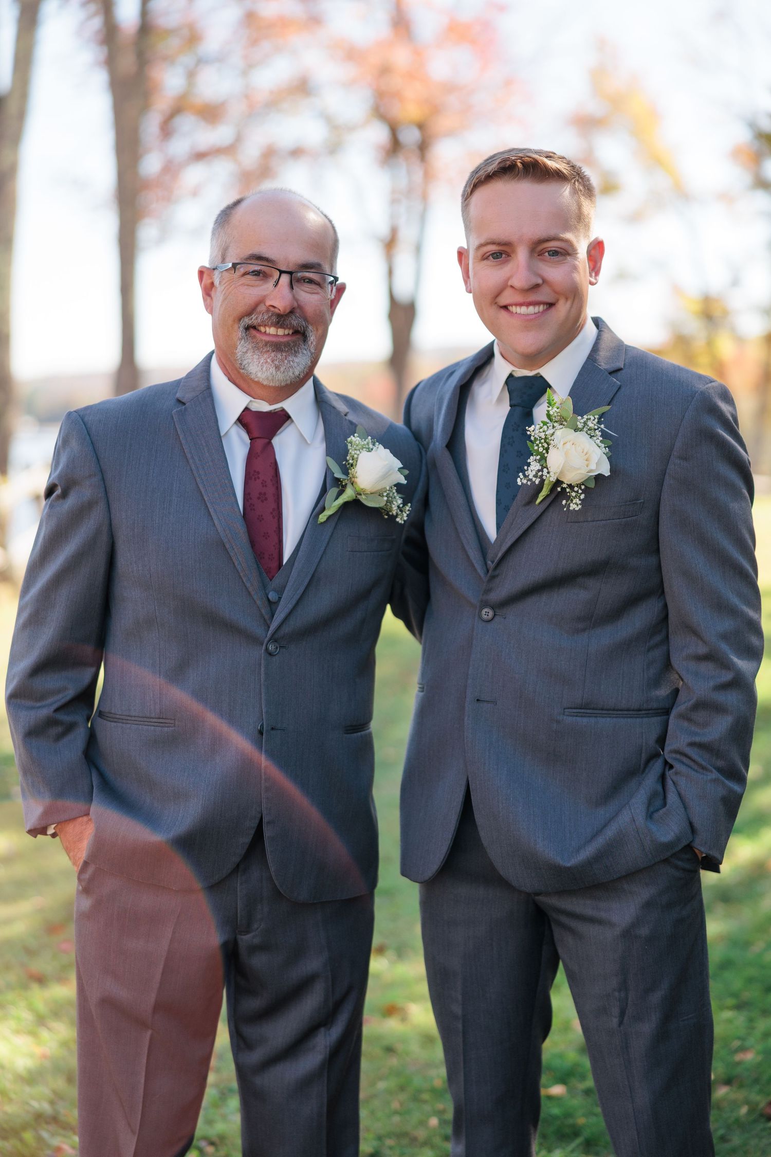 Groomsmen in gray suits with white boutonnieres pose together outdoors during autumn.