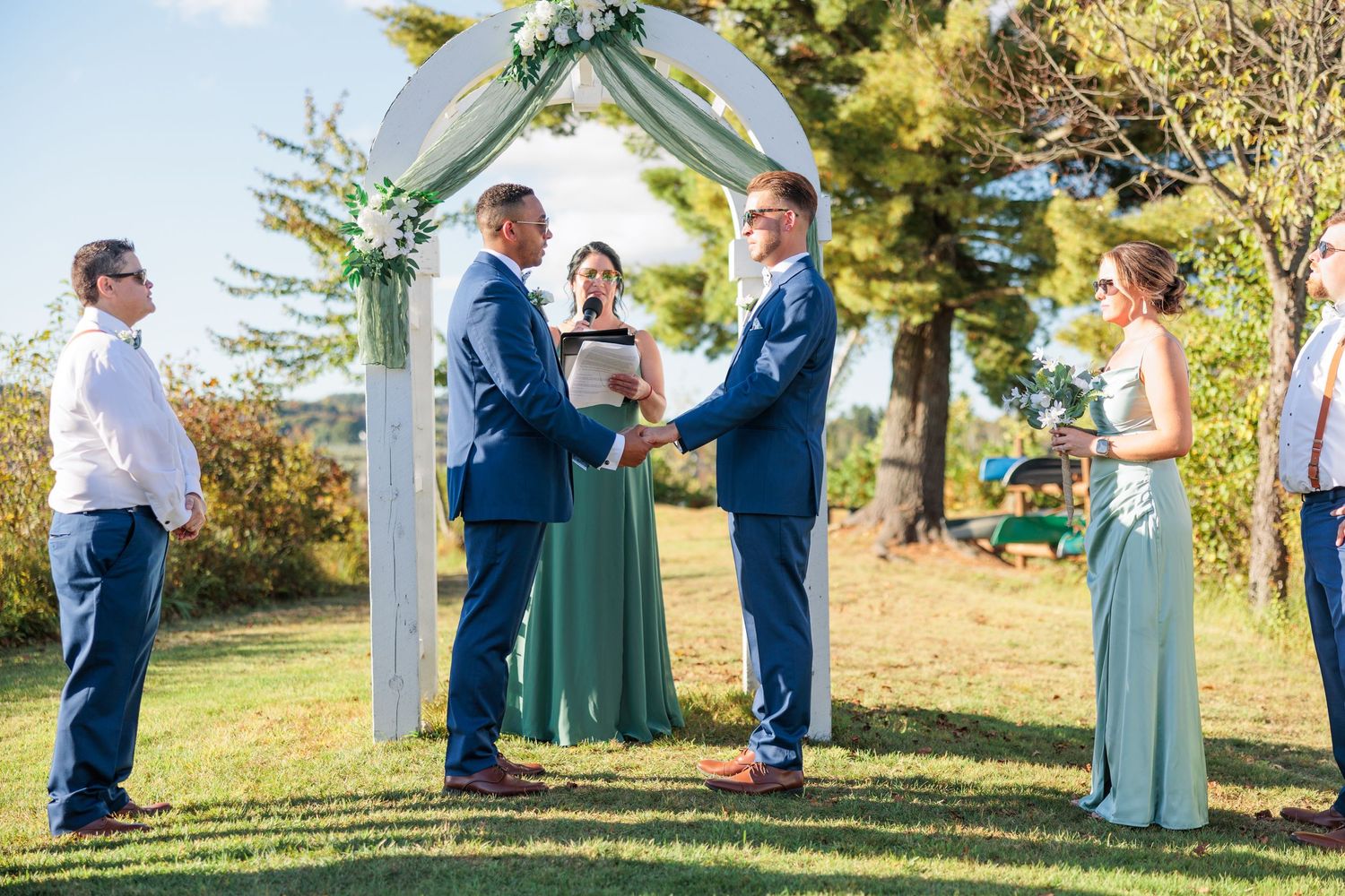 Wedding party members in blue suits and sage green dresses stand under a white ceremony arch.