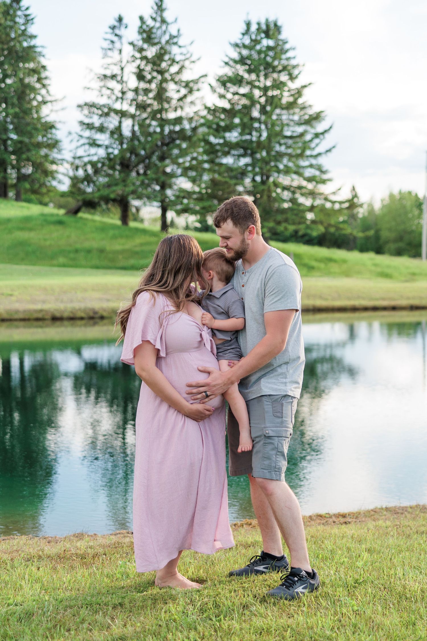 Pregnant woman in pink dress with husband and toddler son posing by a pond surrounded by green trees.