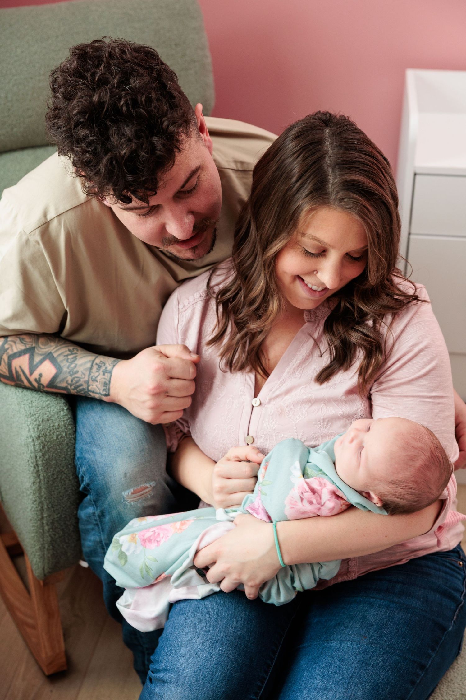 Young couple smiling at their newborn baby wrapped in floral swaddle in a pink nursery room.
