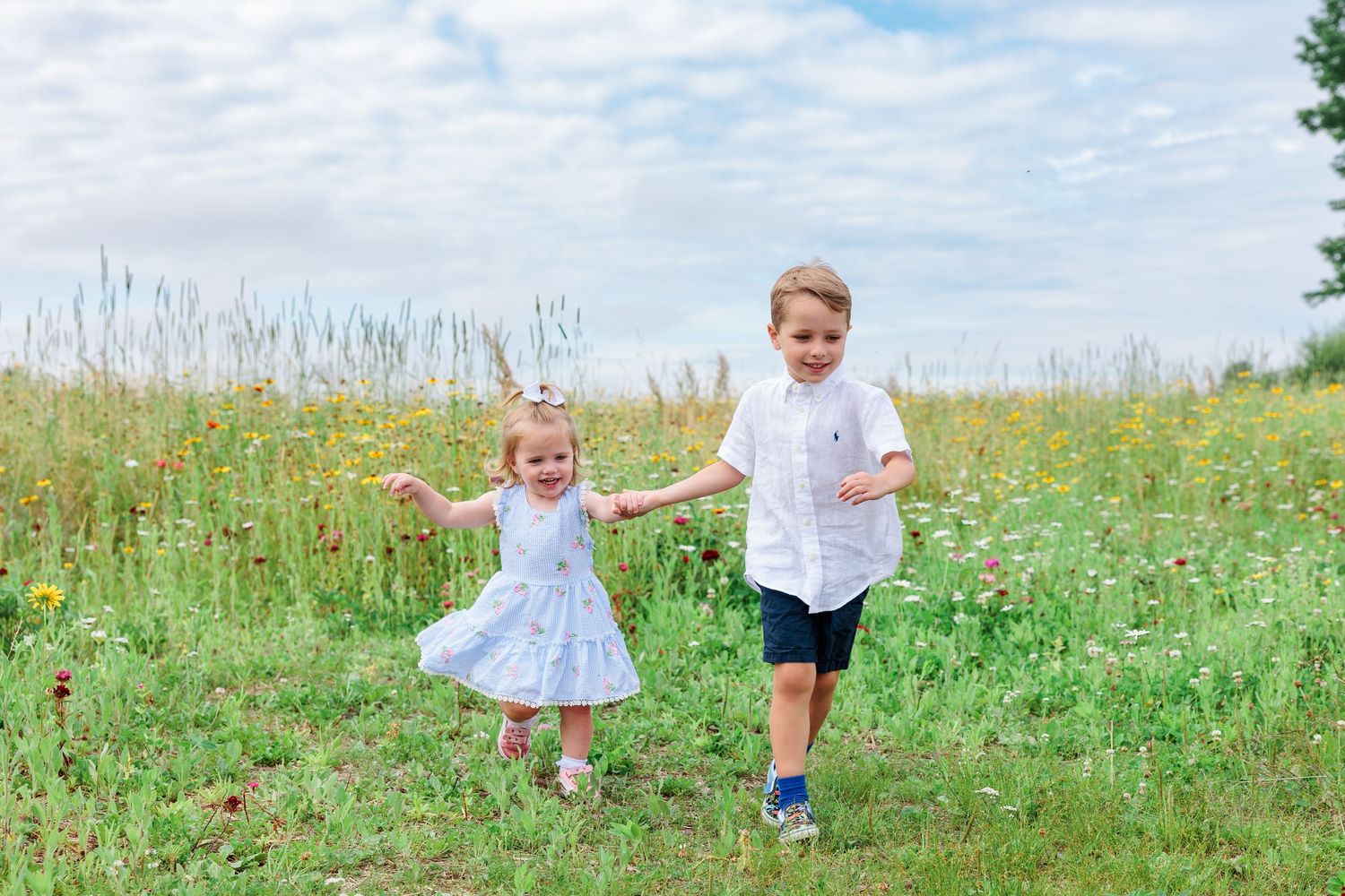 Two young siblings laughing and running together through a colorful summer wildflower meadow.