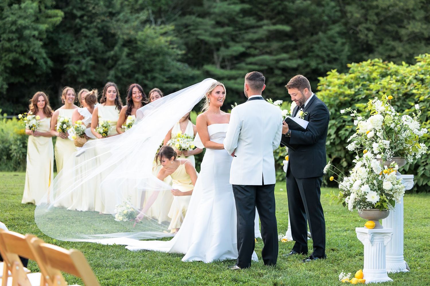Bride in flowing gown with dramatic veil exchanging vows with groom at elegant outdoor garden wedding ceremony.