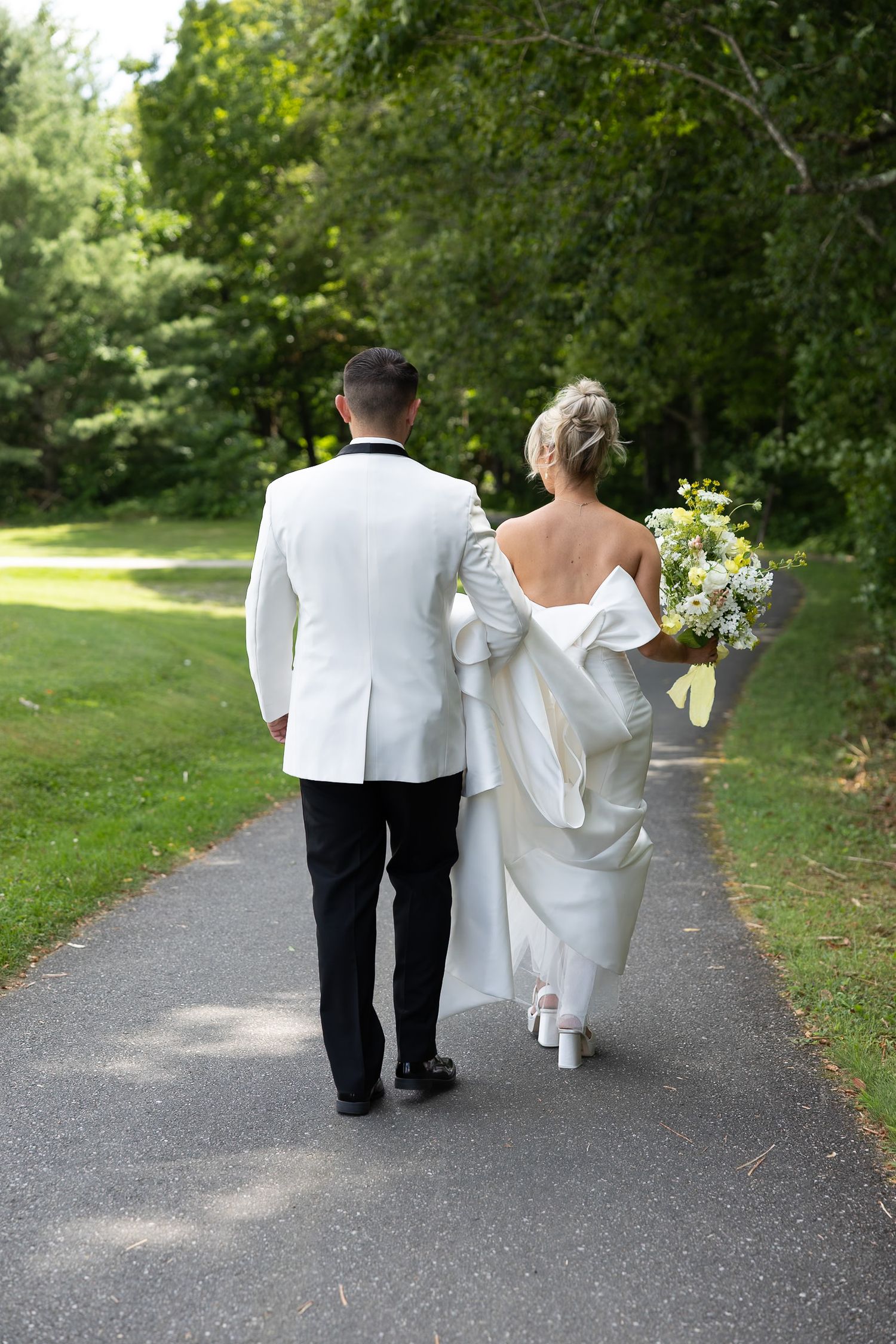 Bride and groom walking hand in hand down a tree-lined path, viewed from behind on their wedding day.