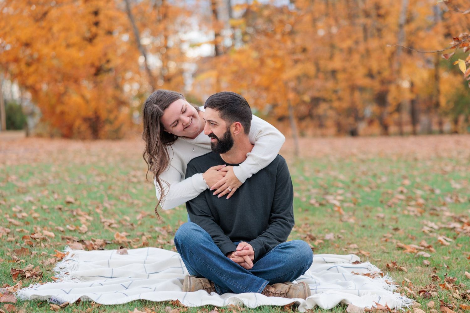 Woman hugging man from behind as he sits cross-legged on blanket in park with golden autumn leaves.
