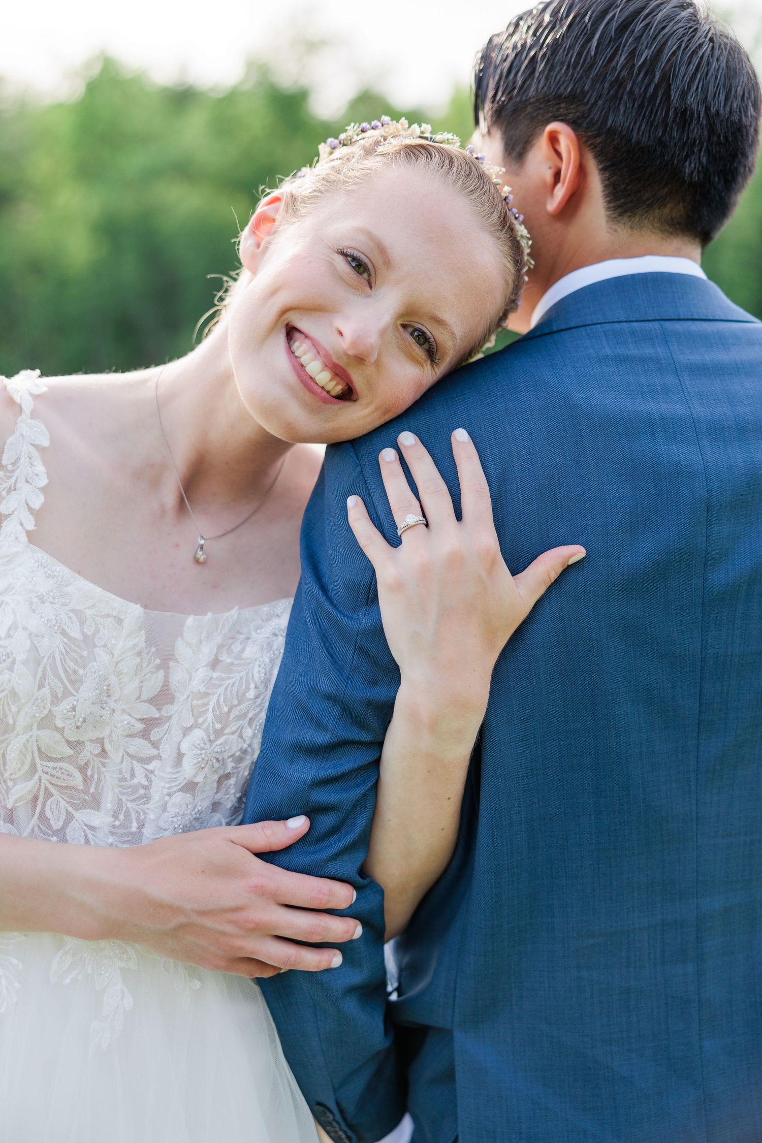 Radiant bride in white lace dress smiling while embracing groom in navy suit outdoors on wedding day.