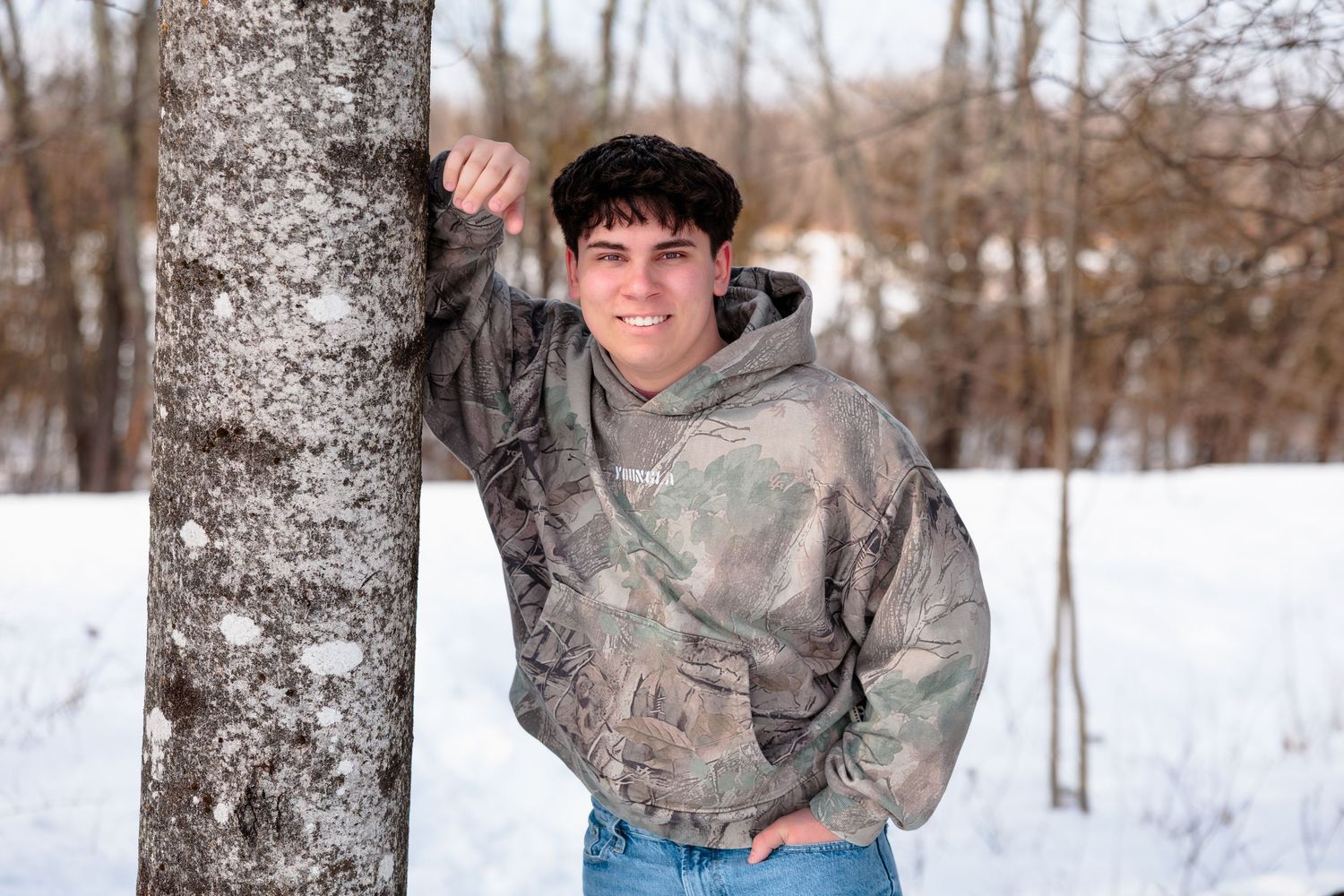 A smiling teenage boy in a camo hoodie leans against a birch tree in a snowy winter landscape.