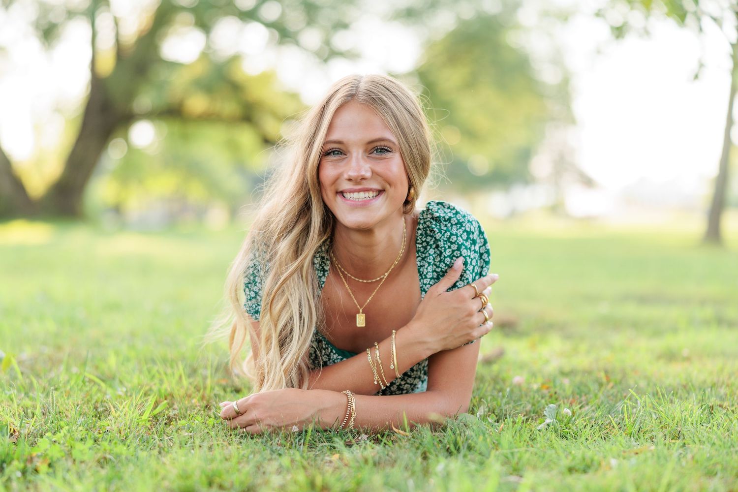 Smiling blonde woman lying on green grass outdoors, wearing a teal floral top and gold necklace, with trees in background.