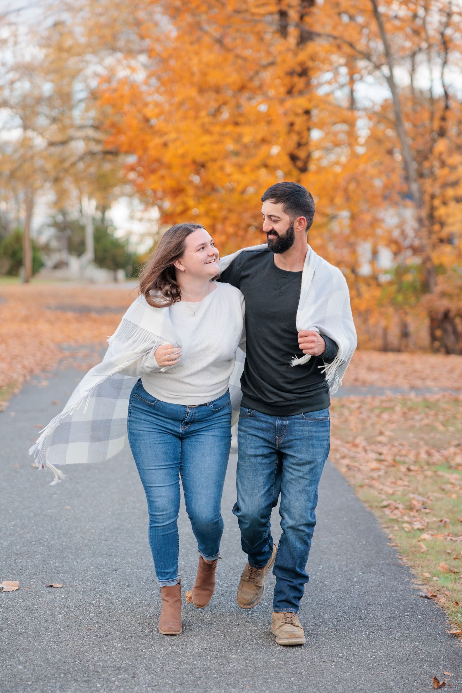 Couple walking together on autumn path sharing a plaid blanket, surrounded by vibrant orange fall foliage.