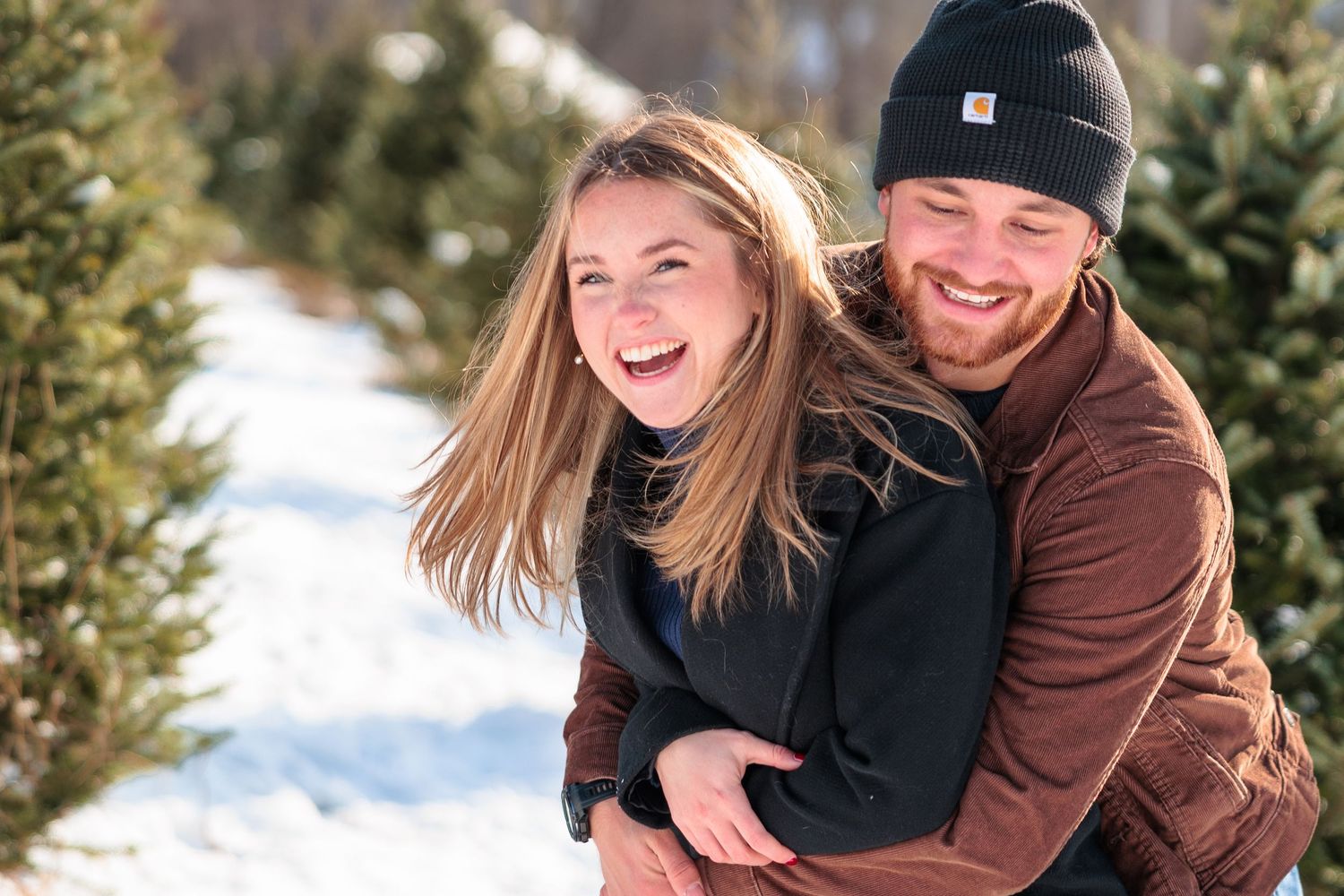 Laughing couple enjoying a piggyback ride at a snowy Christmas tree farm on a sunny day.