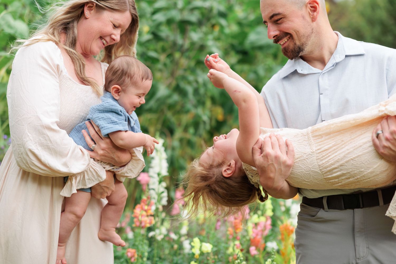 Joyful family laughing as father playfully holds young child upside down in colorful garden.