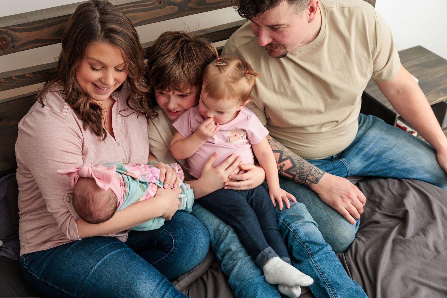 Family of five with newborn baby and two young children sitting closely together on a bed.