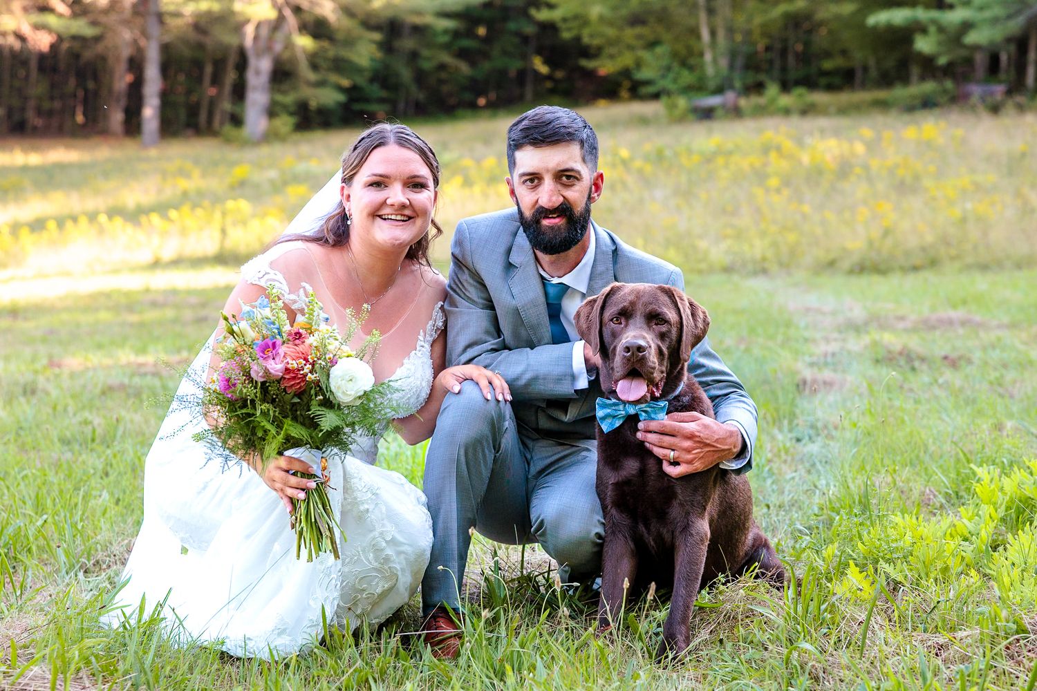 Newlyweds pose with their chocolate Labrador wearing a blue bow tie in a sunny meadow at their wedding.