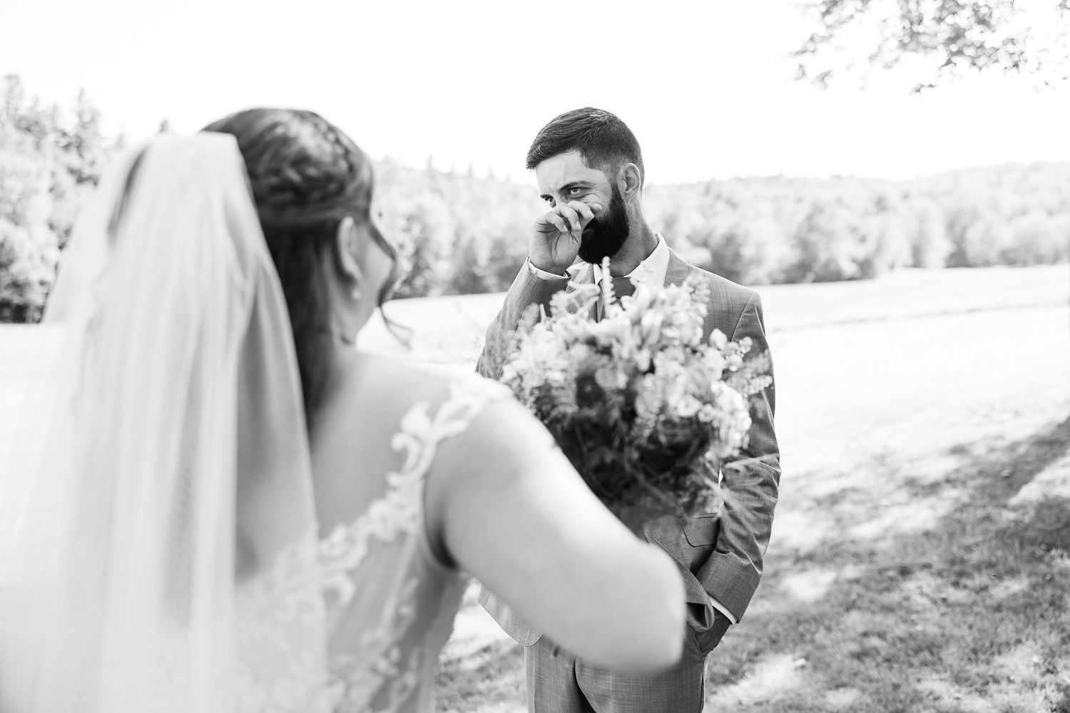 Groom wipes tears of joy during emotional first look with bride outdoors.