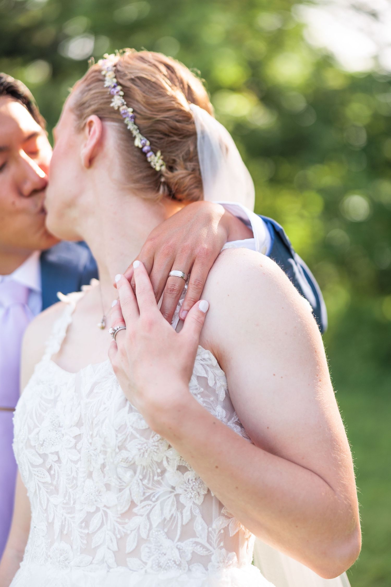 Close-up of bride with floral headpiece and wedding ring embracing groom outdoors in soft natural light.