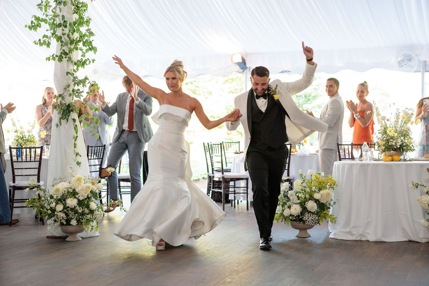 Bride and groom joyfully entering wedding reception under white tent while guests celebrate and applaud around them.