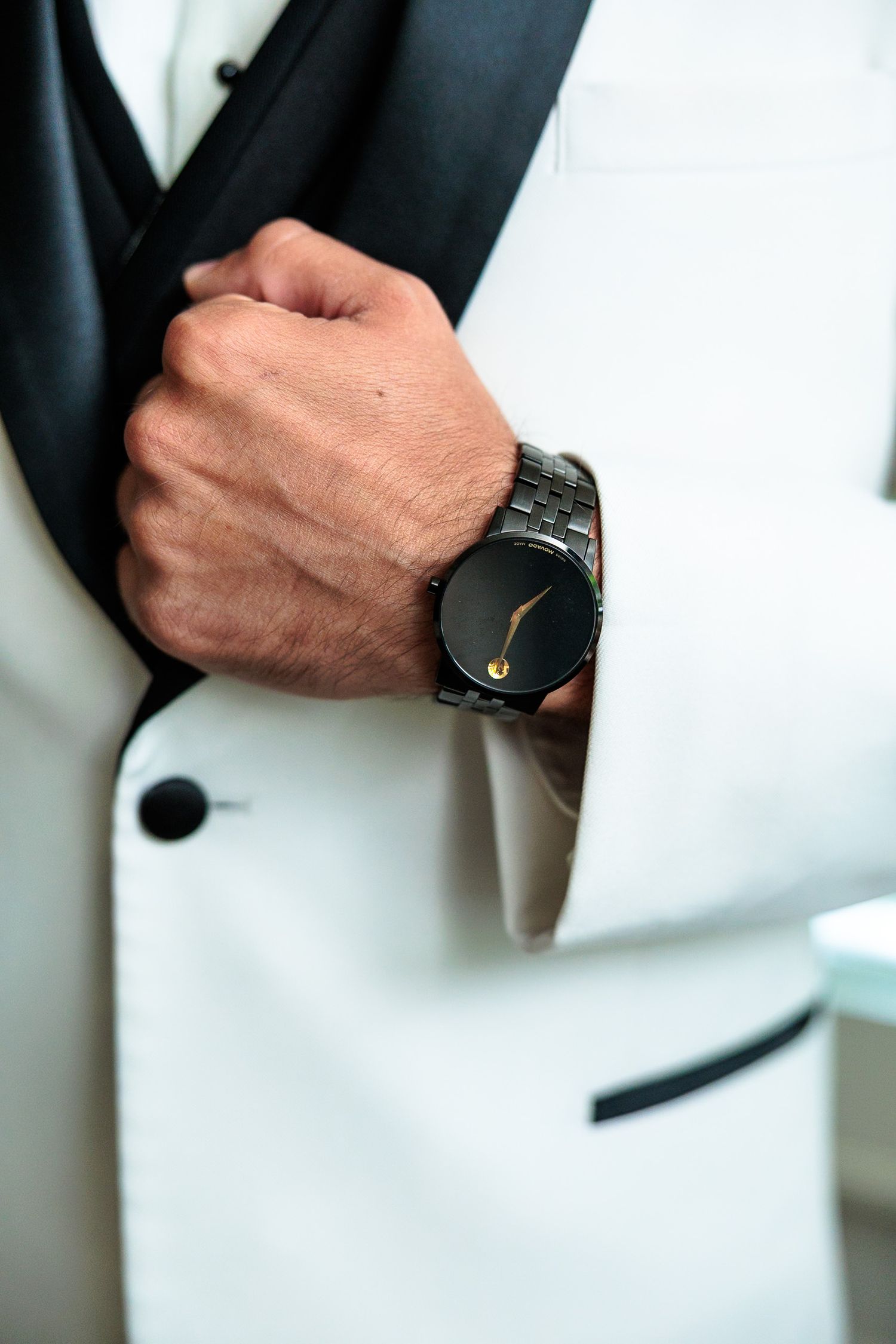 Groom wearing a white tuxedo jacket with a black Movado watch on his wrist at a wedding.