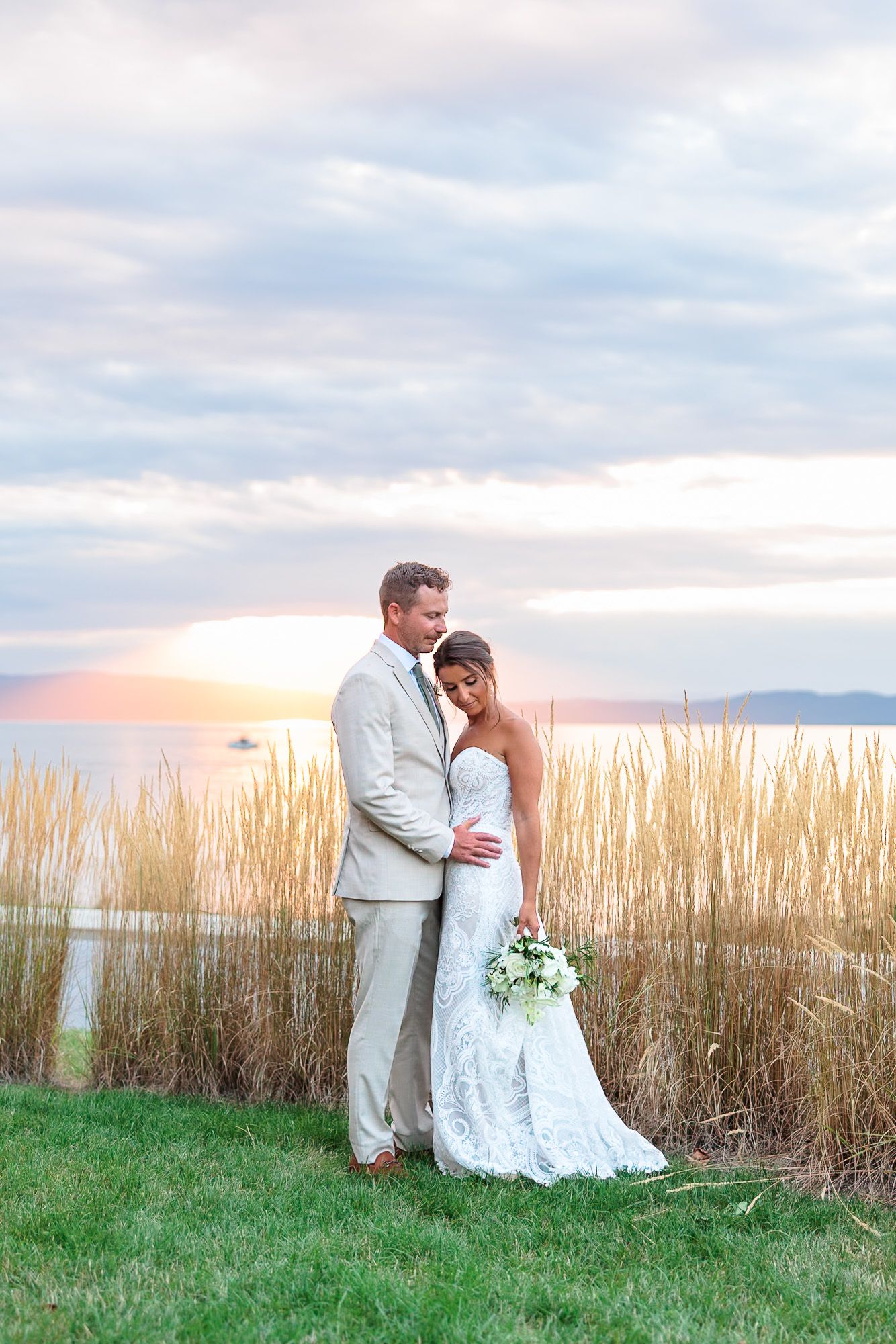 Bride and groom embrace at sunset by tall golden reeds near a lake, bride holding white bouquet in lace gown.