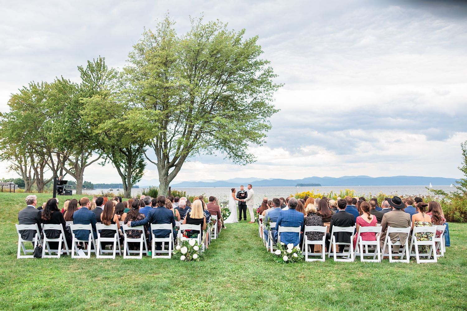 Outdoor wedding ceremony on green lawn by waterfront with guests seated in white chairs under large trees.