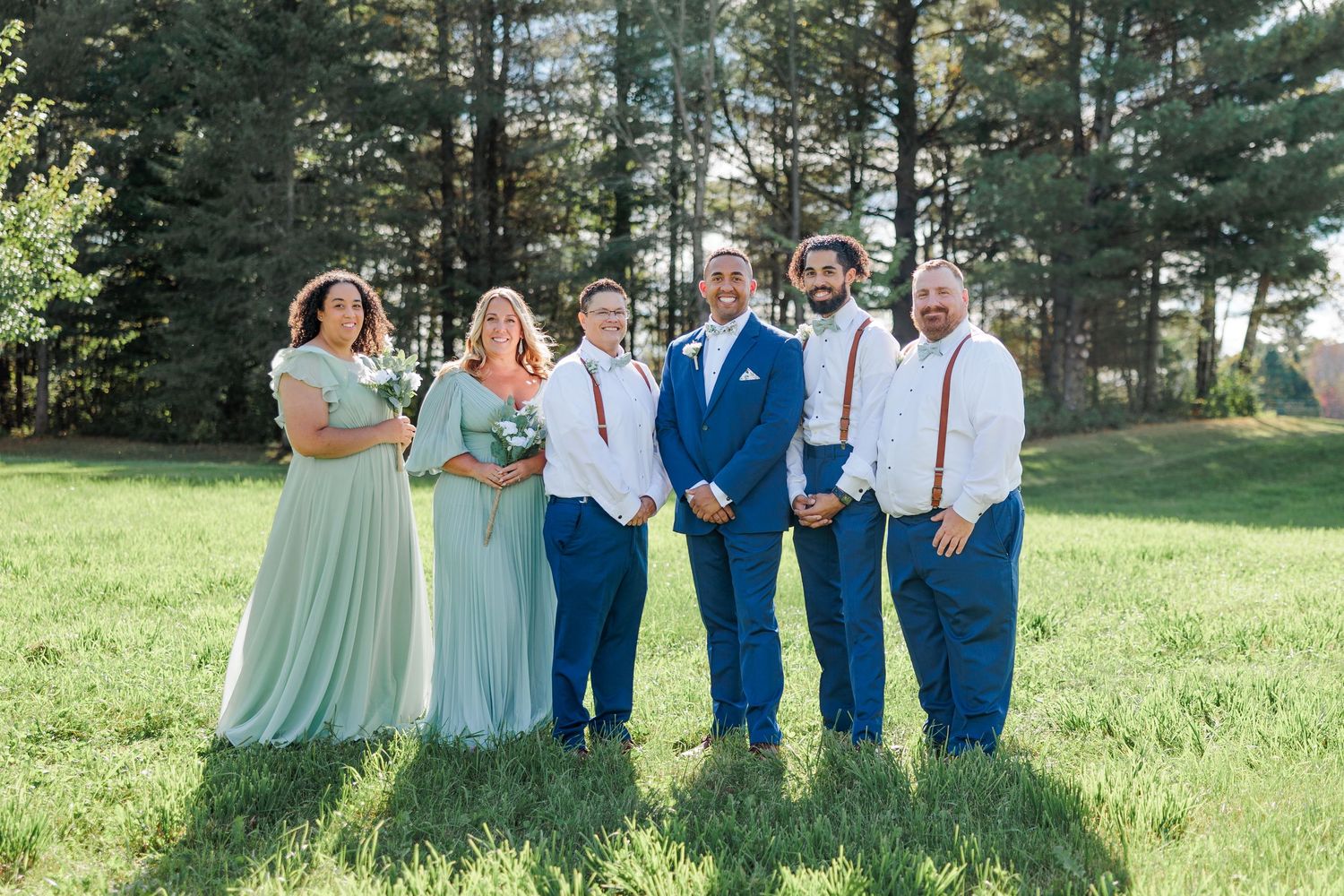 Wedding party in mint green dresses and navy suits pose together on sunny lawn with pine trees in background.