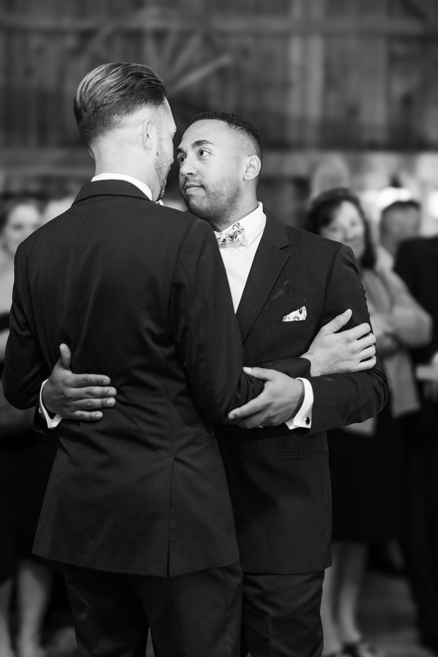 Two people in black suits share an intimate dance moment at a formal event.