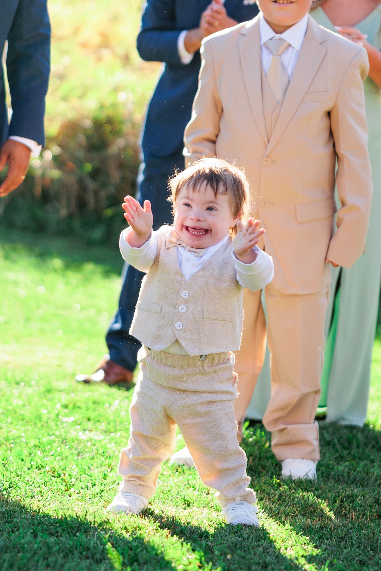 A happy toddler in a beige suit laughs while playing on grass during a wedding celebration.