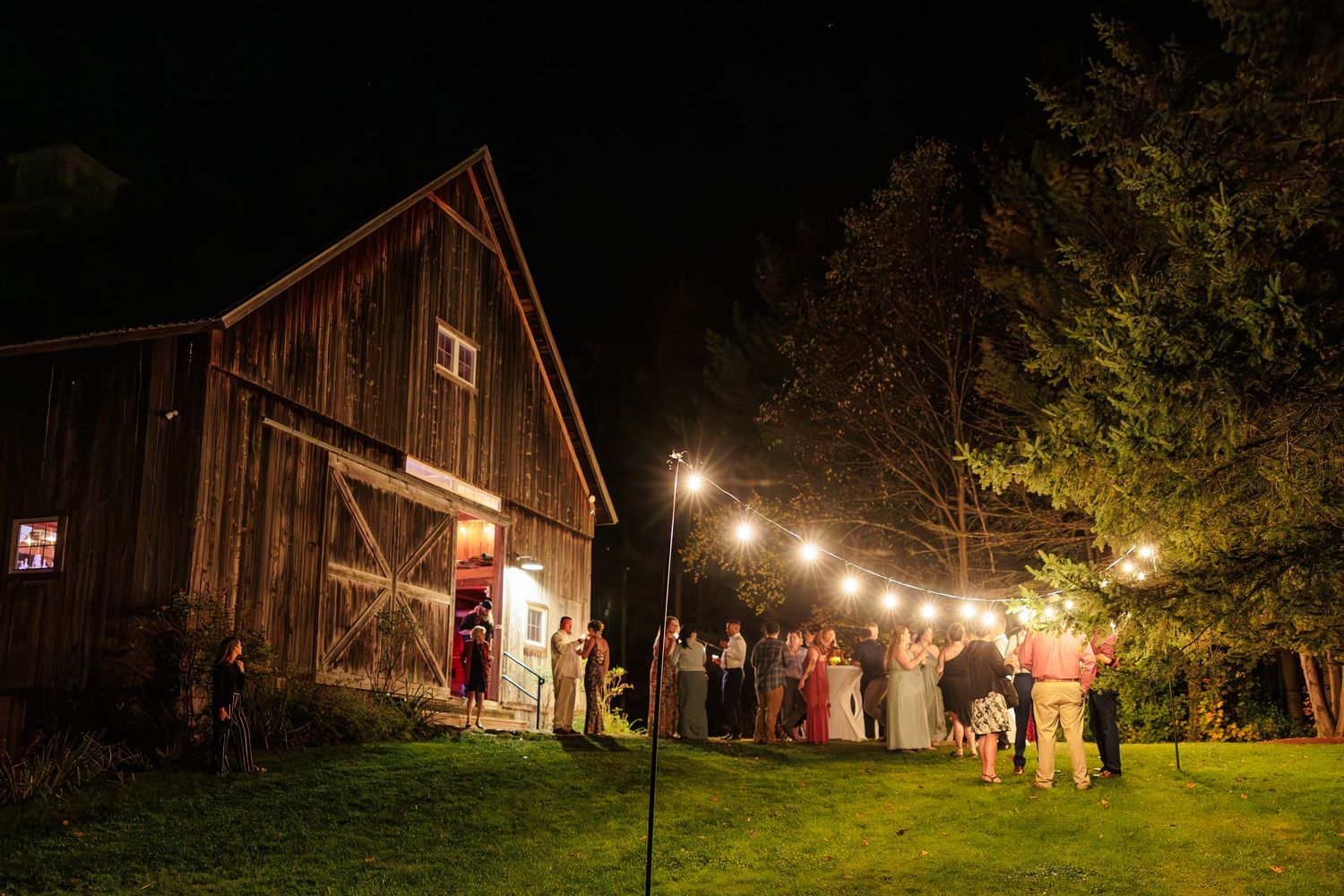 A rustic barn wedding venue illuminated by string lights at night with guests gathered outside.