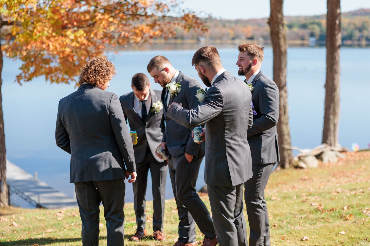A group of groomsmen gather by a lakeside on an autumn day wearing matching gray suits.