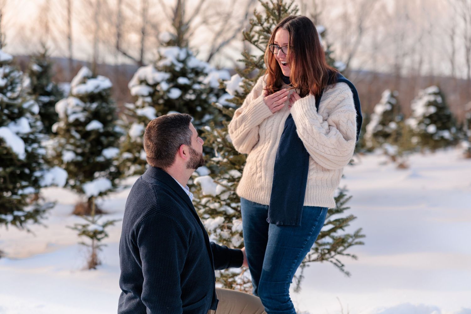A couple shares a romantic moment during a winter engagement photoshoot among snow-covered evergreen trees at sunset.
