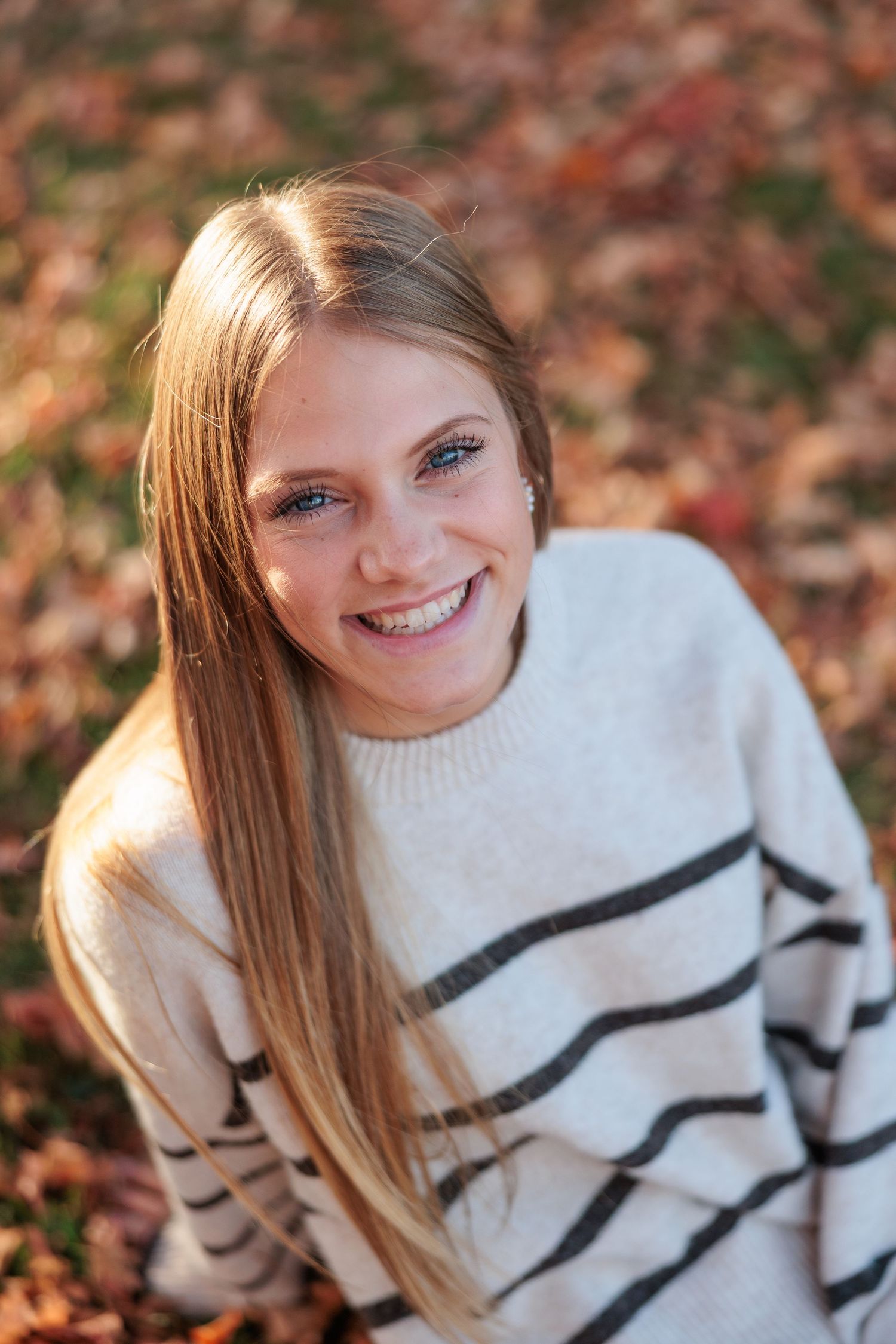 Smiling teenage girl with long blonde hair wearing a striped sweater sitting among fall leaves outdoors.