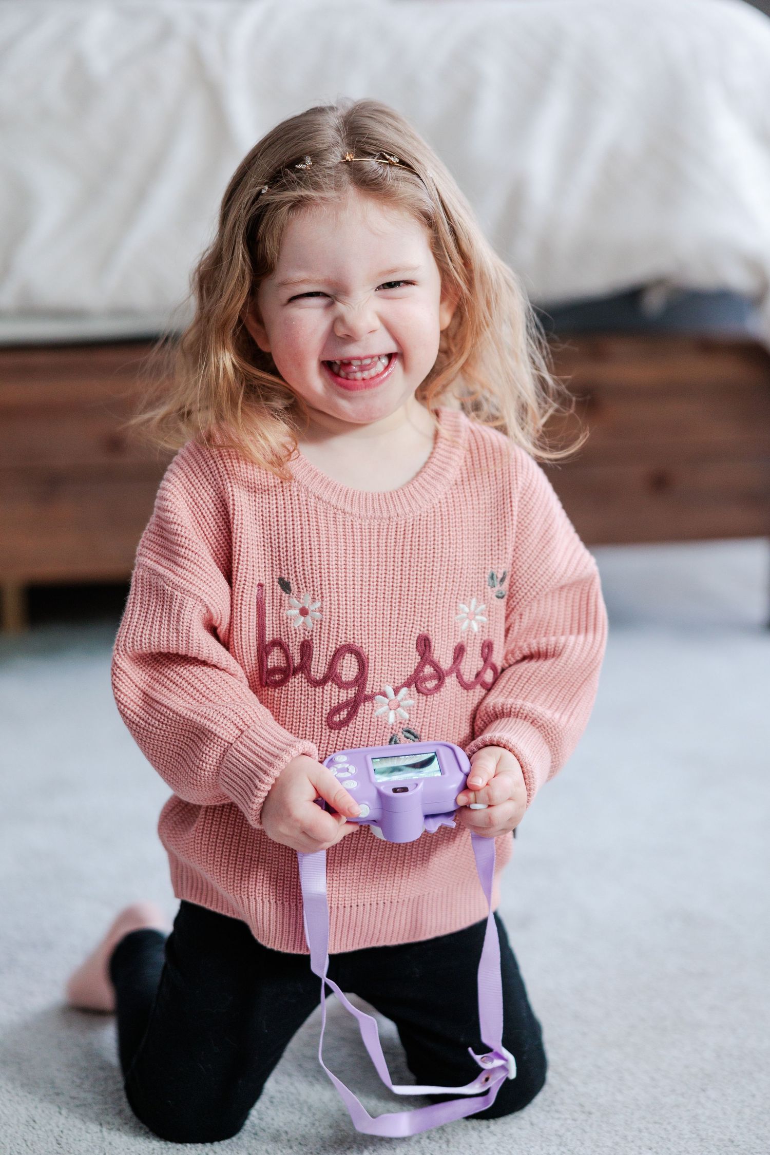 Happy toddler girl in pink big sis sweater laughing while holding a toy camera.