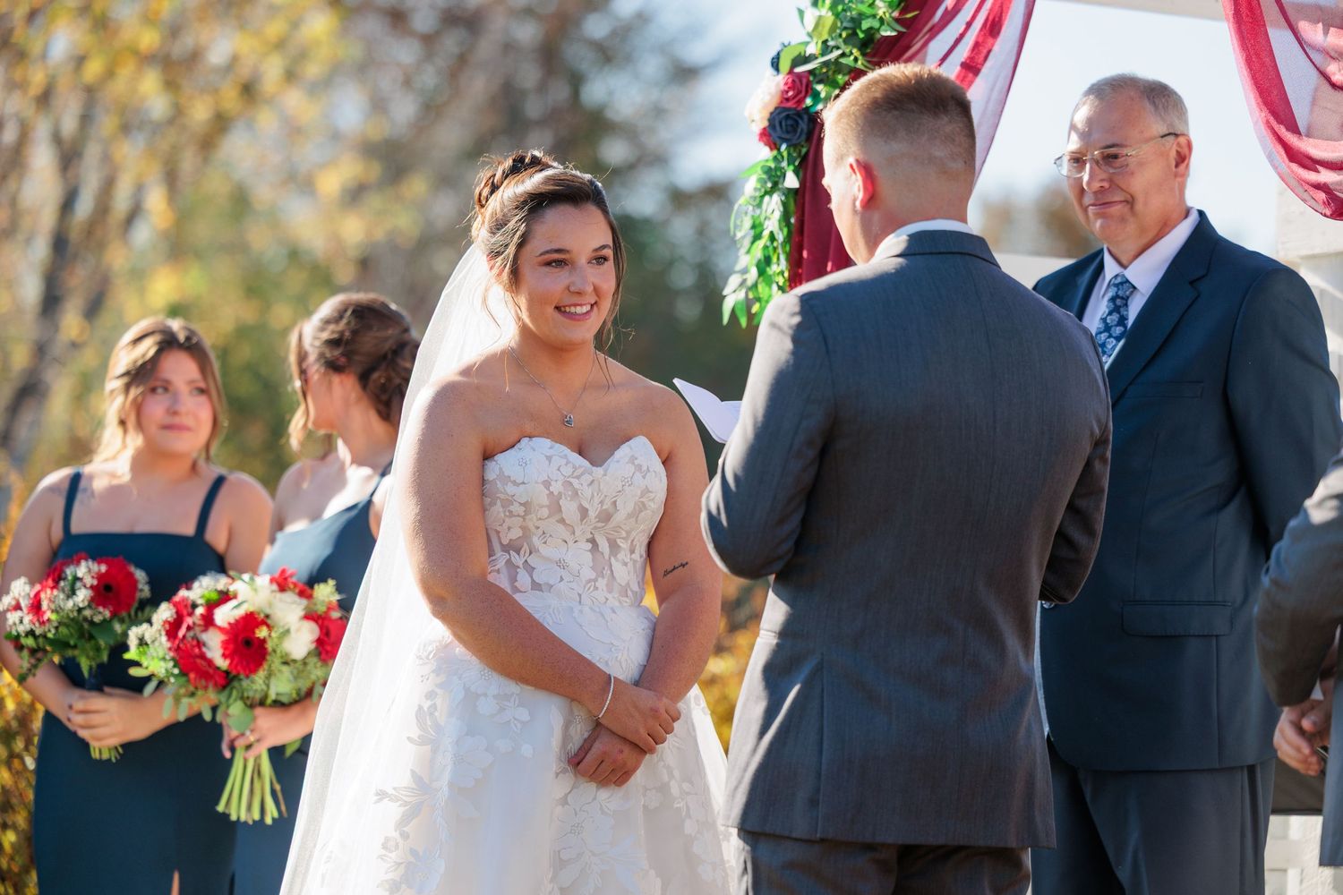 Smiling bride in strapless lace gown faces groom at outdoor fall wedding ceremony with floral arch.