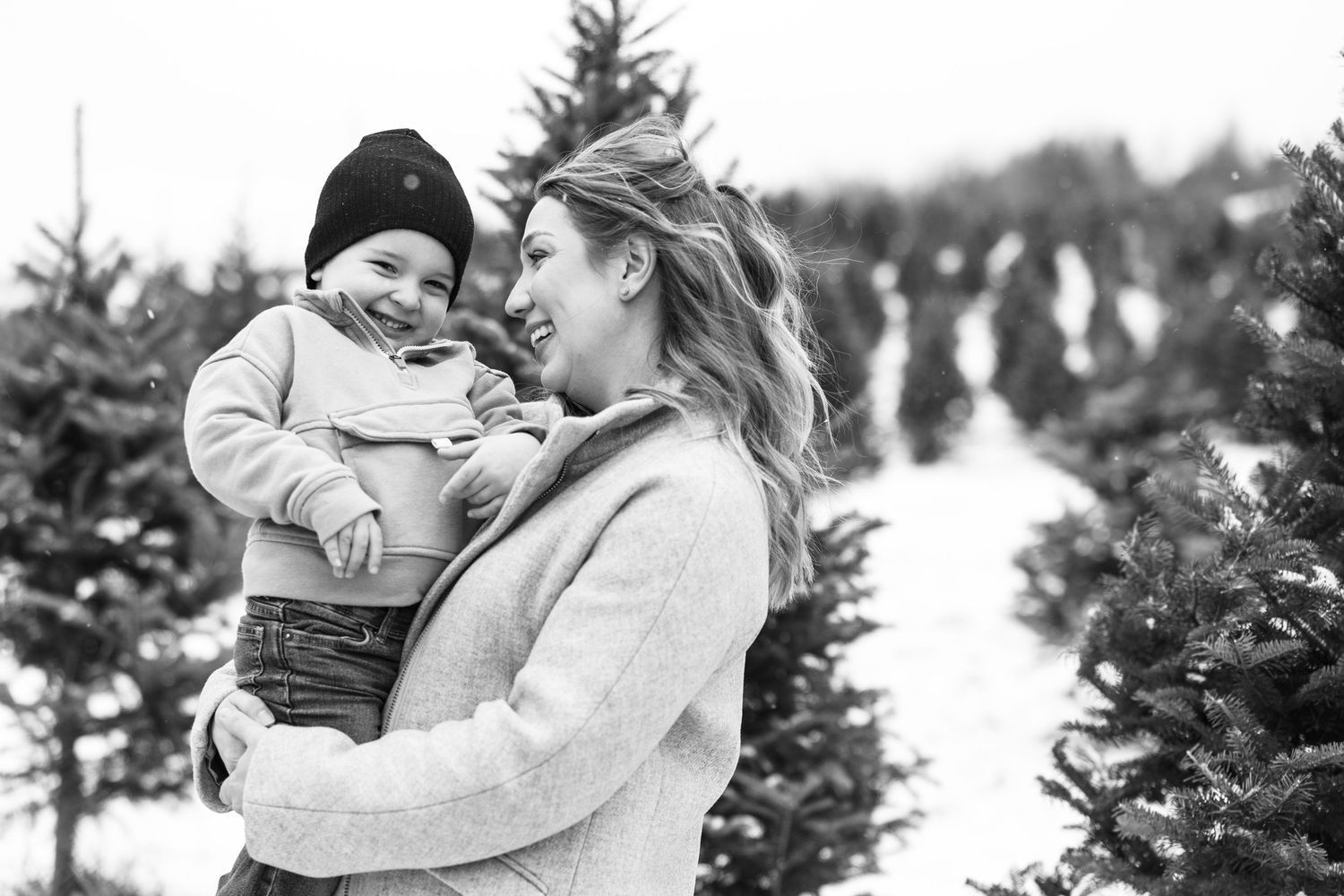 Black and white photo of a smiling mother holding her happy toddler at a Christmas tree farm.
