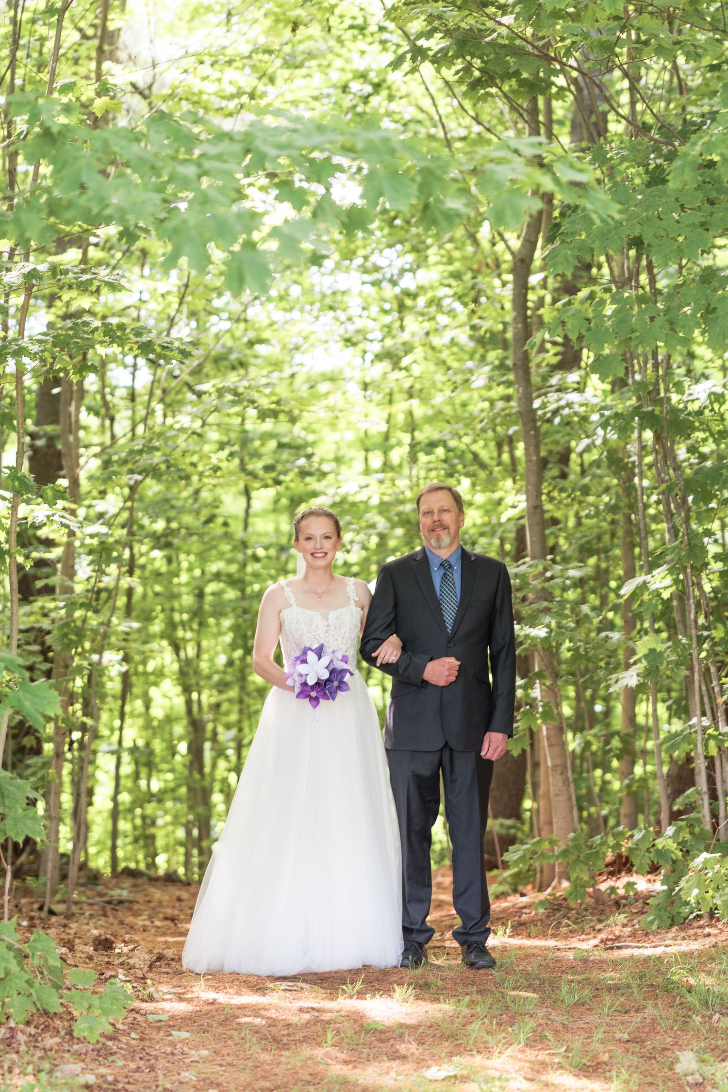 Smiling bride in white gown holding purple bouquet walking with father through a sunlit forest path.