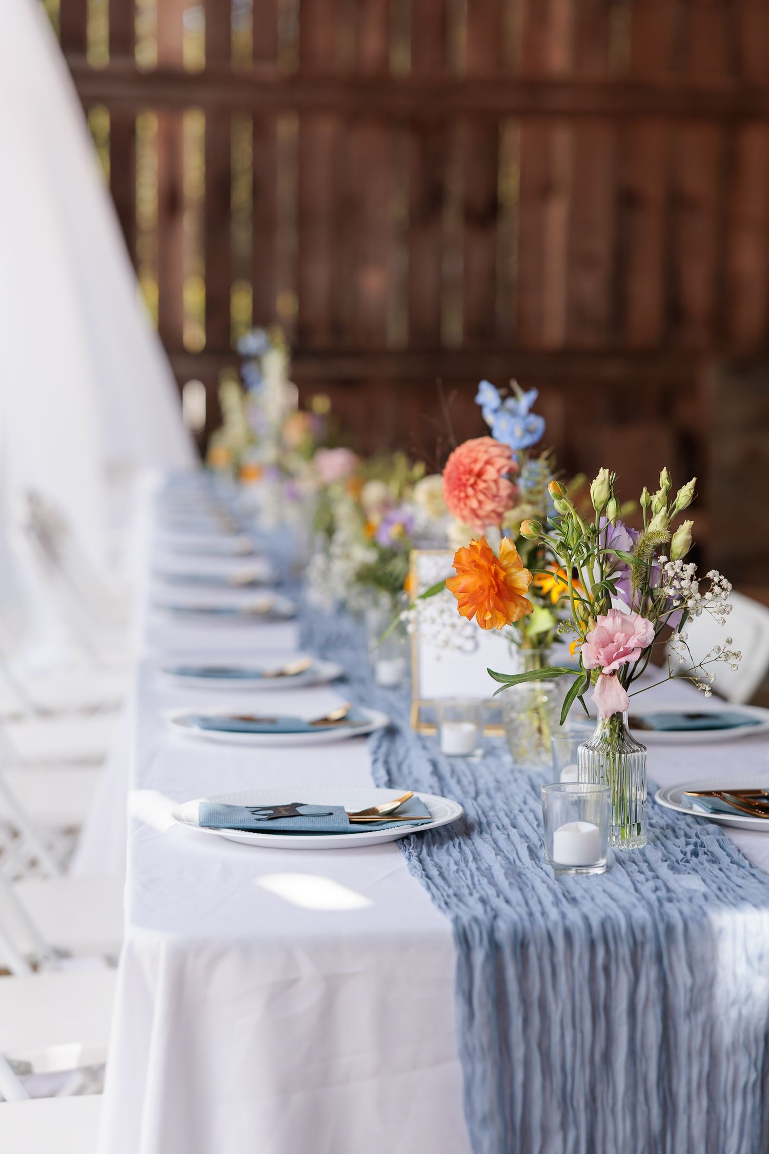 Elegant wedding reception table with dusty blue runner, colorful wildflower centerpieces, and blue charger plates in a rustic barn.