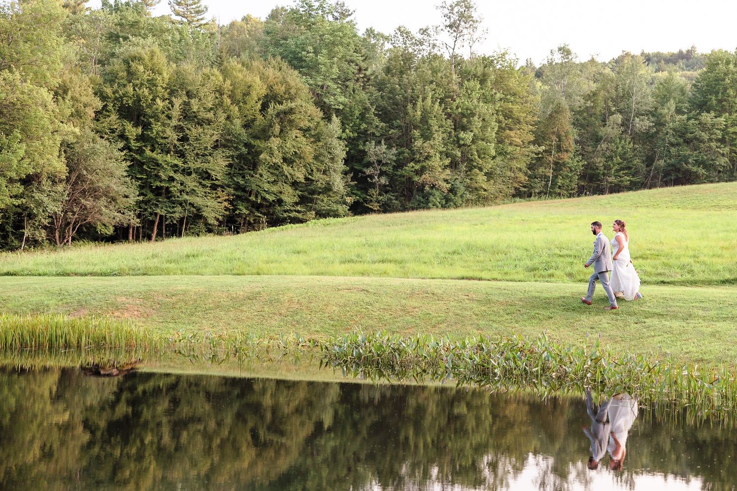 Bride and groom walking hand in hand along a lush green pond reflection landscape at golden hour.