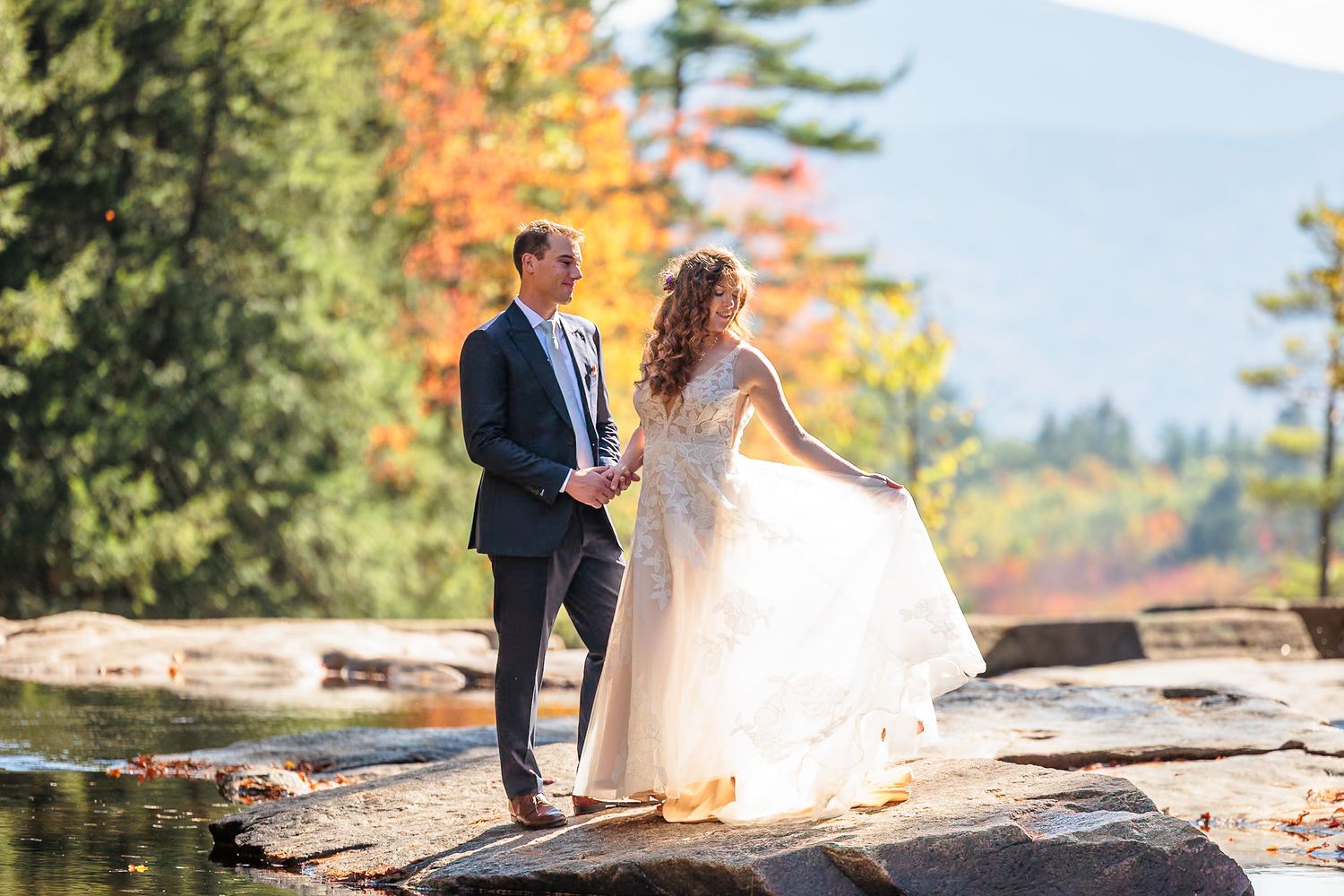Bride and groom hold hands on rocky riverbank surrounded by vibrant autumn foliage and mountain backdrop.
