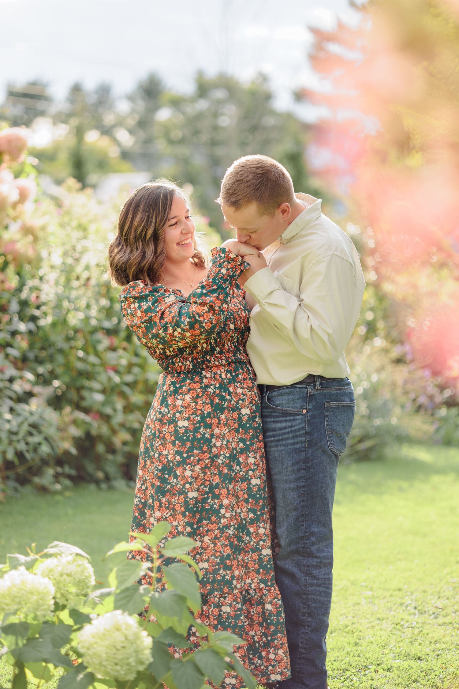 Man kissing womans hand as she smiles in a lush garden with blooming flowers and soft sunlight.