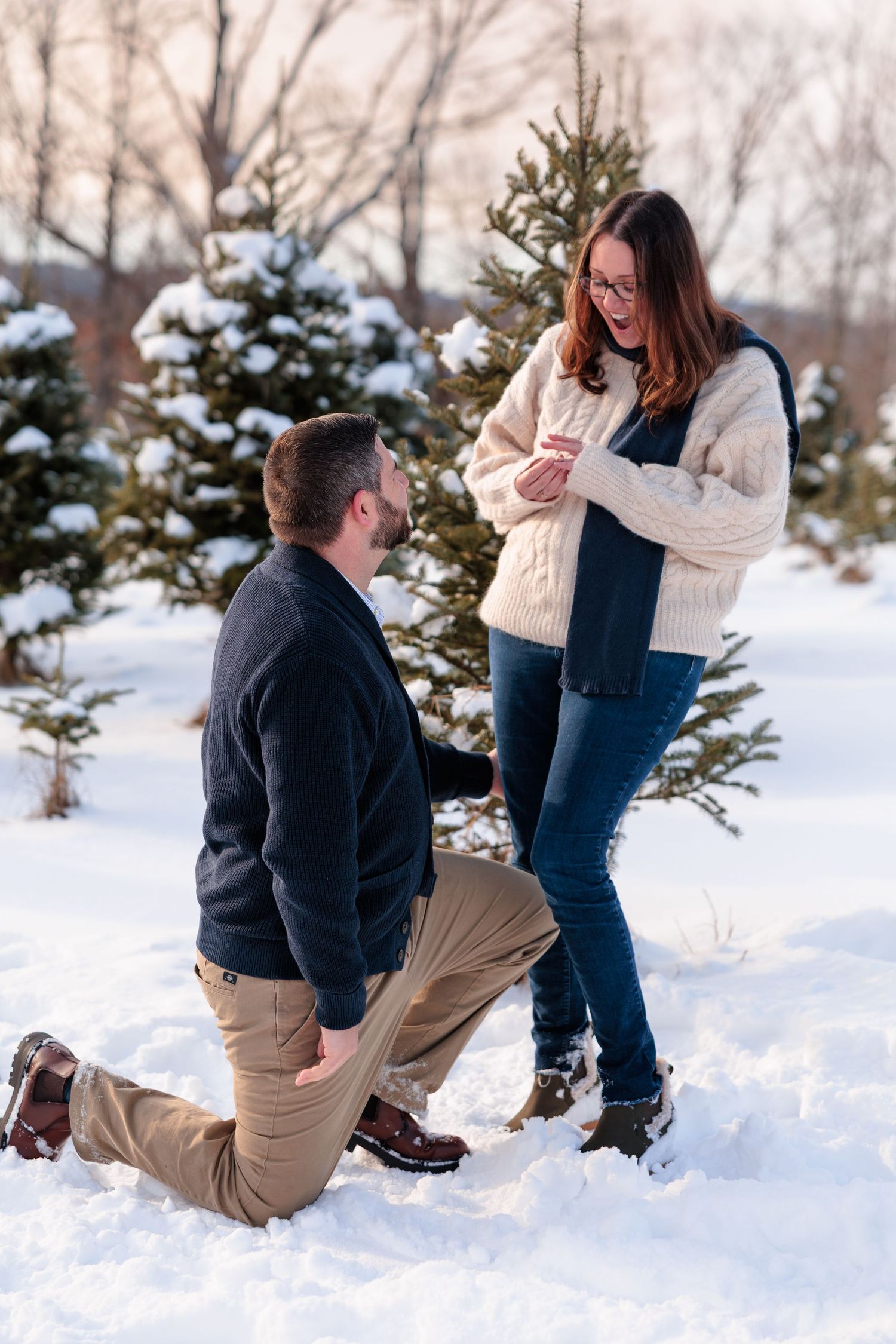 Man kneeling in snow holding woman's hand during a winter marriage proposal at tree farm.