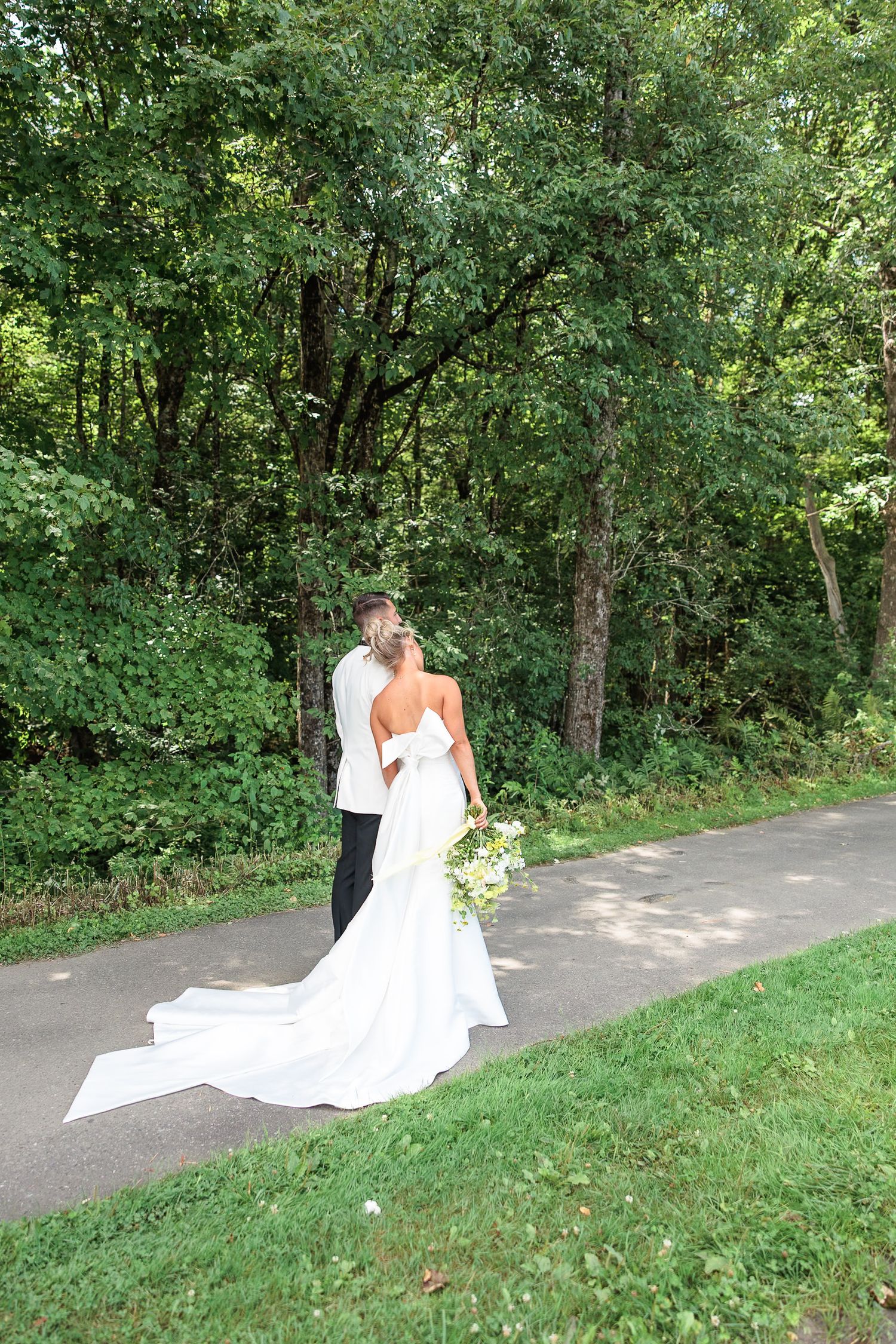Bride in strapless gown with long veil and groom walk along a tree-lined path at outdoor wedding.