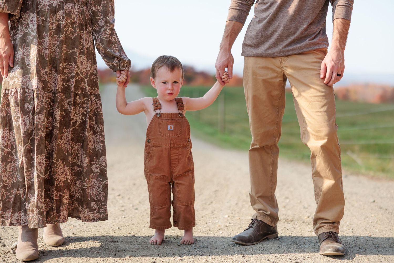 Toddler in brown overalls holding parents hands while standing on a sunny gravel country road.