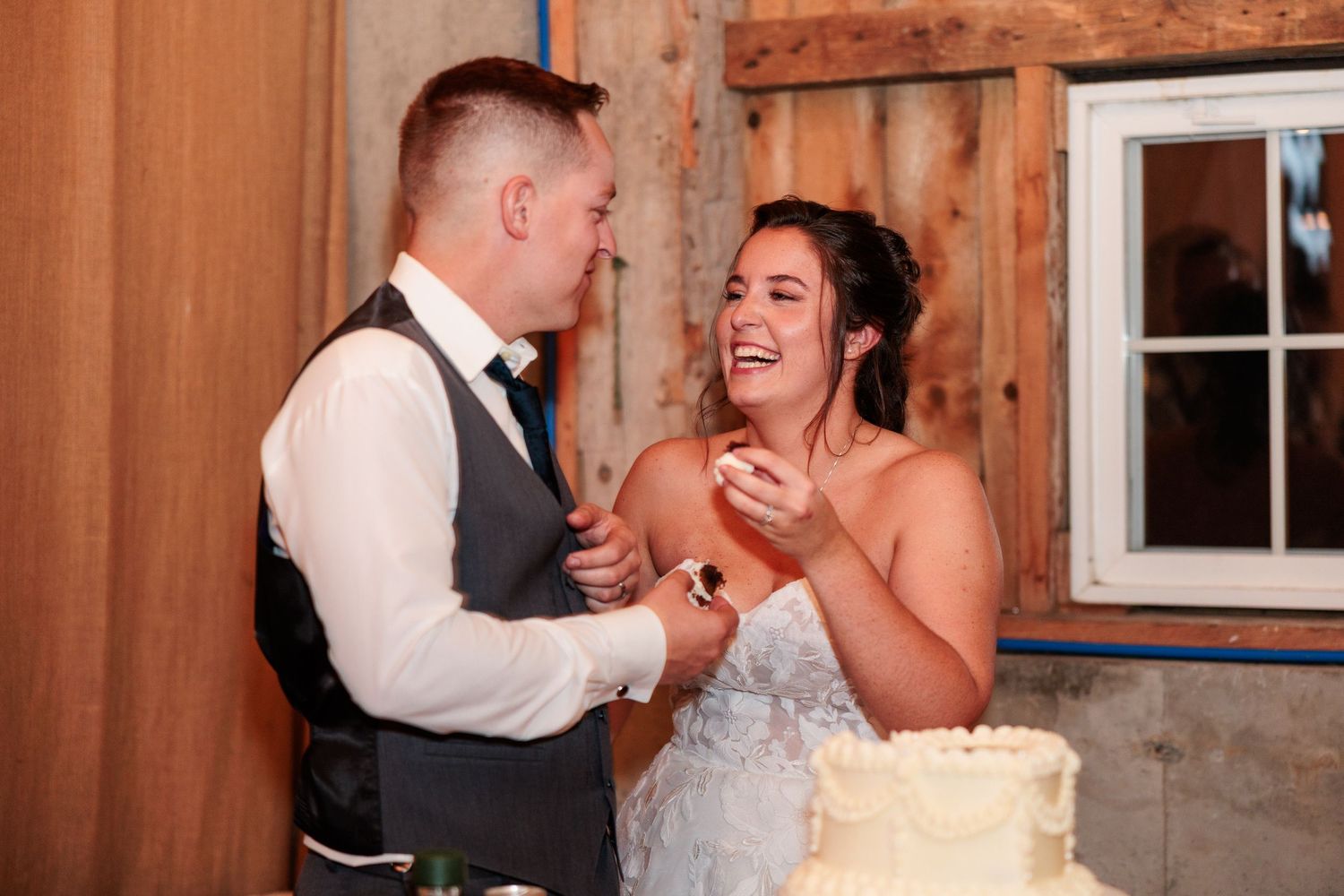 Bride and groom laughing together while cutting their white wedding cake at a rustic barn reception.