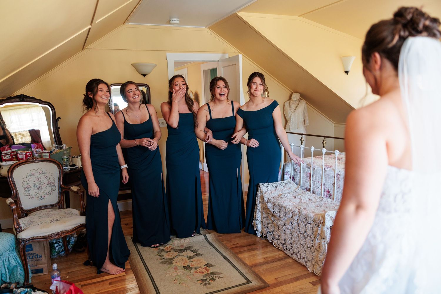 Bridesmaids in navy blue dresses reacting with joy seeing bride in her wedding dress.