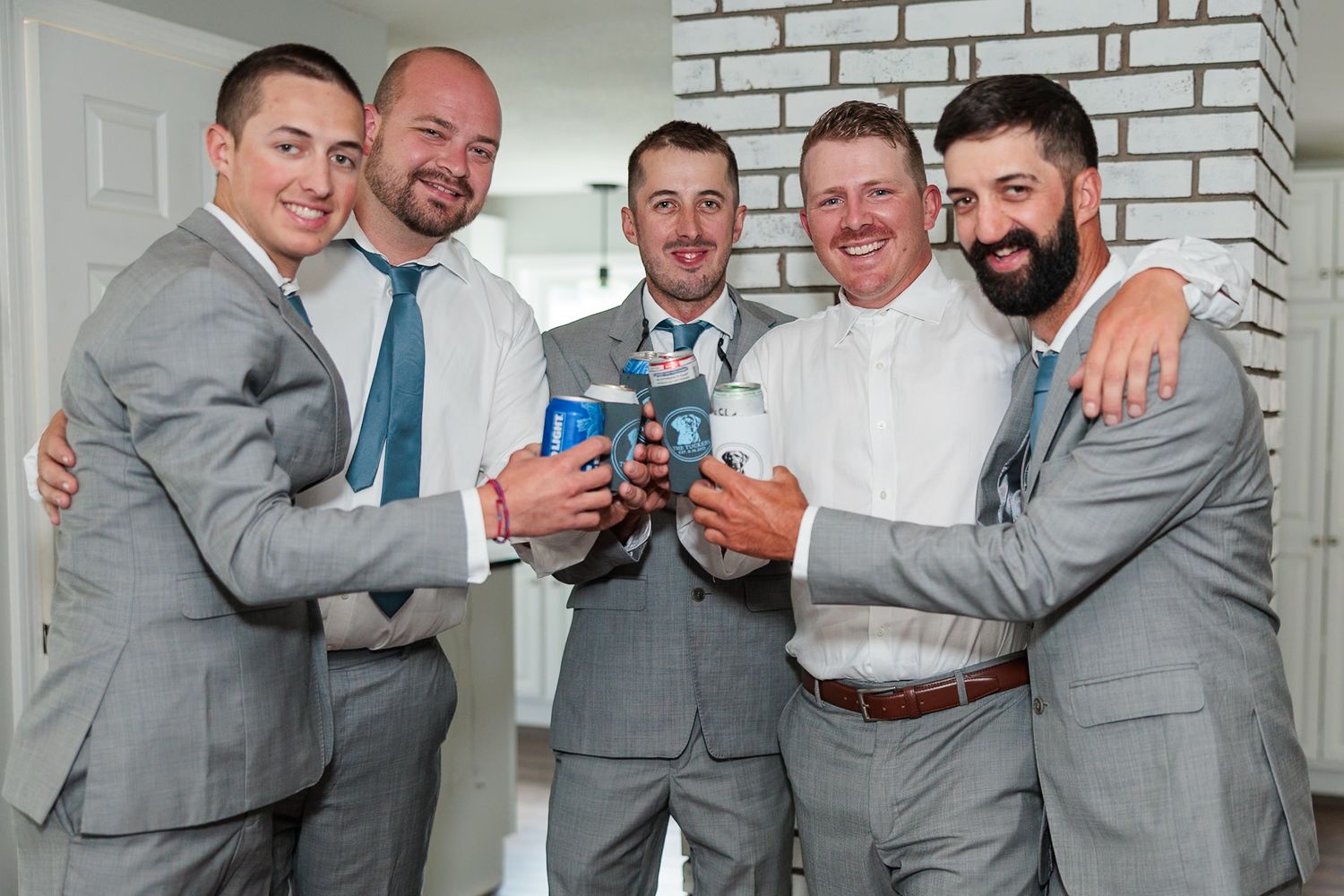 Five groomsmen in gray suits celebrating with drinks and smiling together before wedding ceremony.