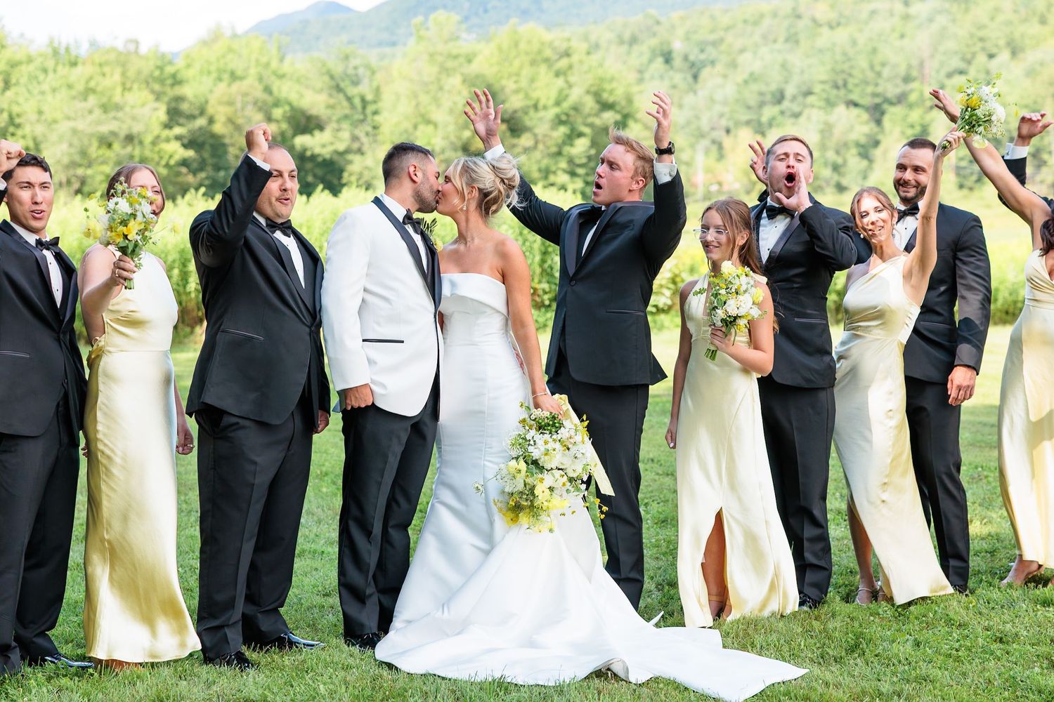 Wedding party celebrates outdoors with bridesmaids in yellow gowns and groomsmen in black tuxedos cheering.