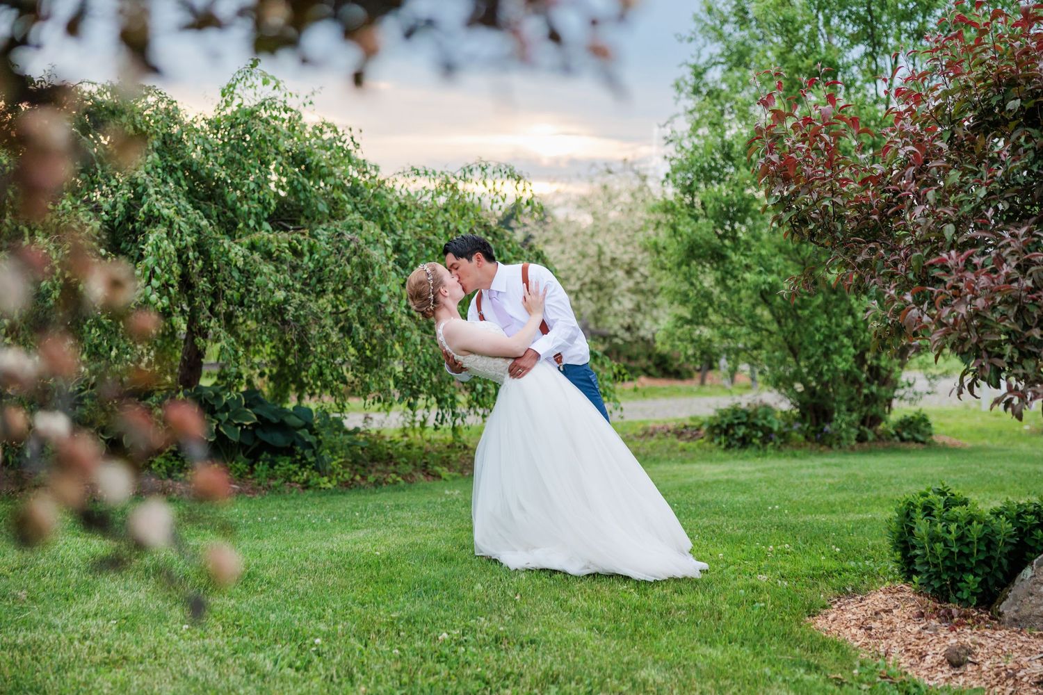 Groom dips bride for a romantic kiss among flowering trees during outdoor wedding portrait session.