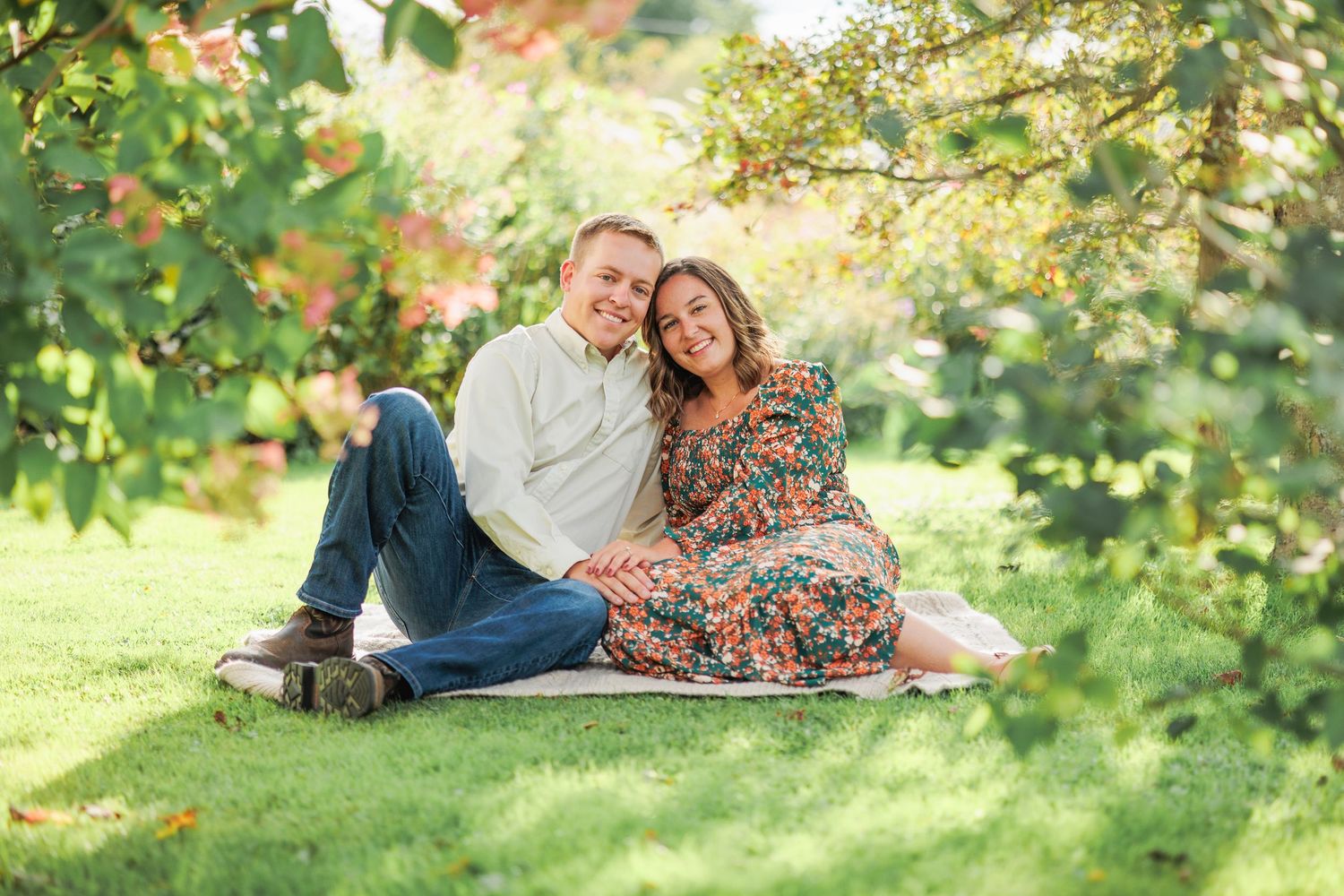 Happy couple sitting together on grass under leafy trees during outdoor engagement photo session.