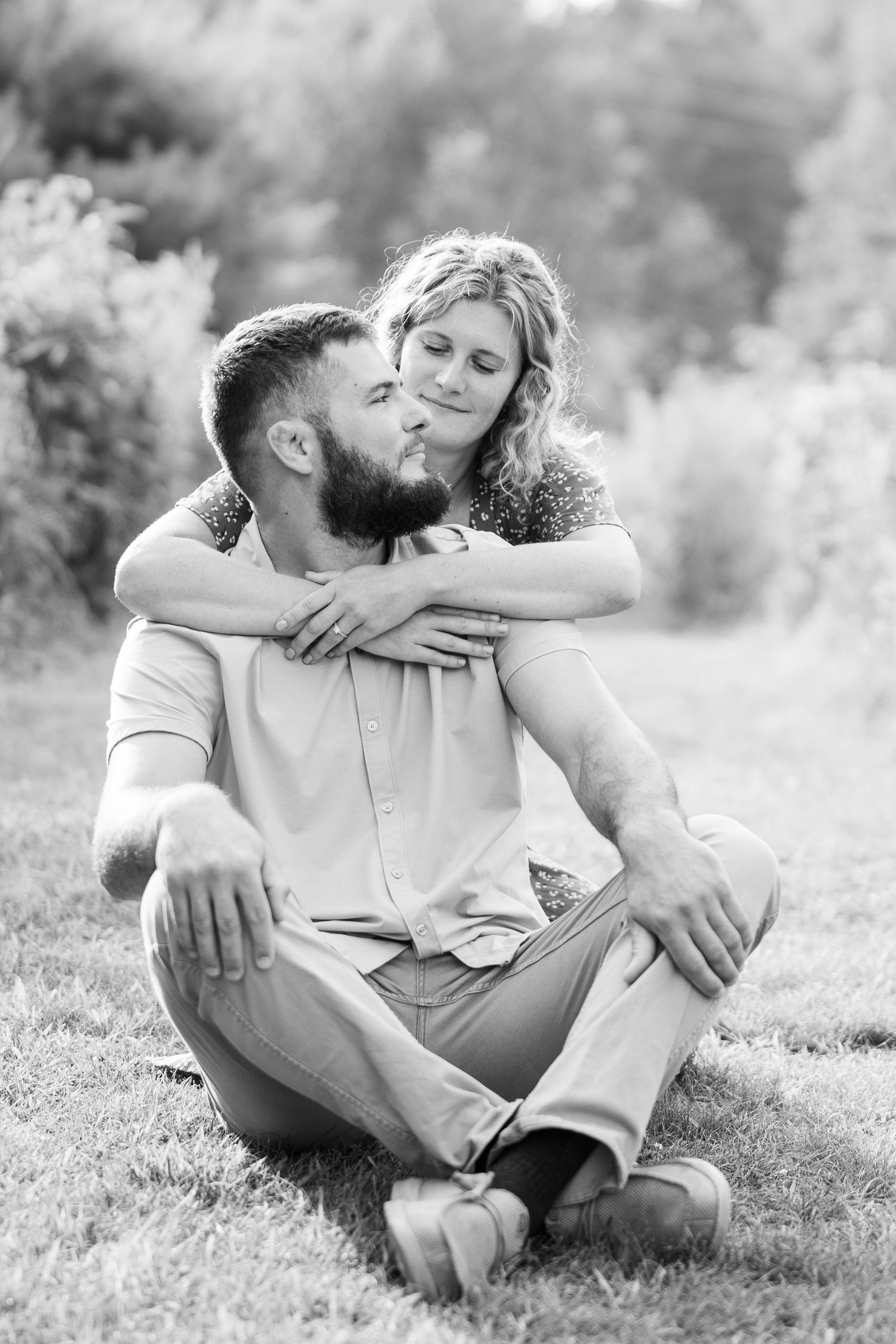 Black and white photo of bearded man sitting on grass while woman embraces him from behind lovingly.