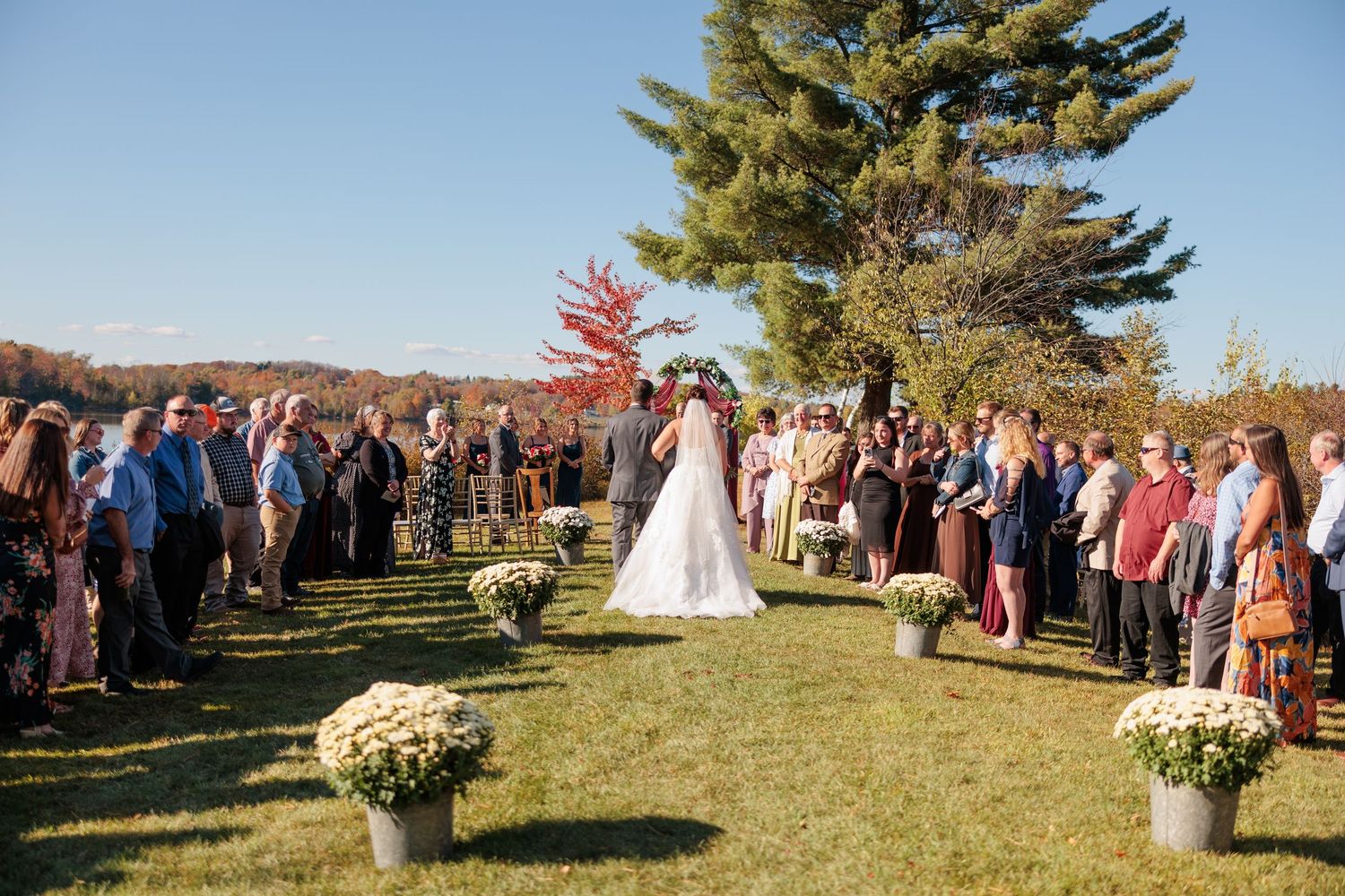 Outdoor fall wedding ceremony with guests lining the aisle as bride walks toward altar.